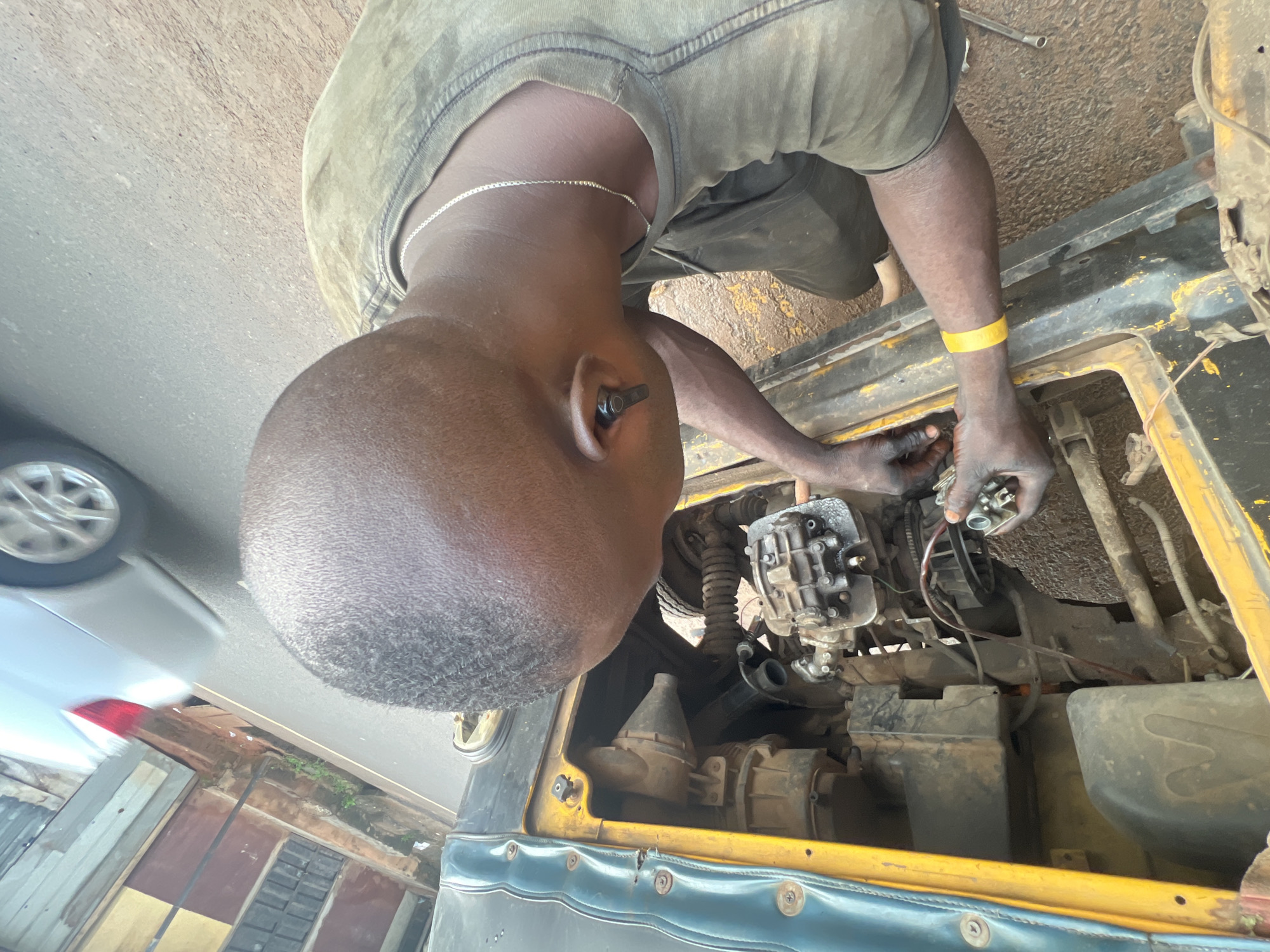 A young African male with short hair, wearing a green t-shirt, dark pants, a silver chain, and an earbud, is shown from the shoulders up, working on the engine of a yellow and blue three-wheeled vehicle. His hands, covered in grease, are visible as he holds a car part. A bright yellow wristband is on his left arm. The vehicle's engine is exposed, with various mechanical components and wires visible. The scene appears to be on a paved road in Nkpor, Nigeria, with buildings and a silver car in the background. The ground is dusty and appears to be a roadside.