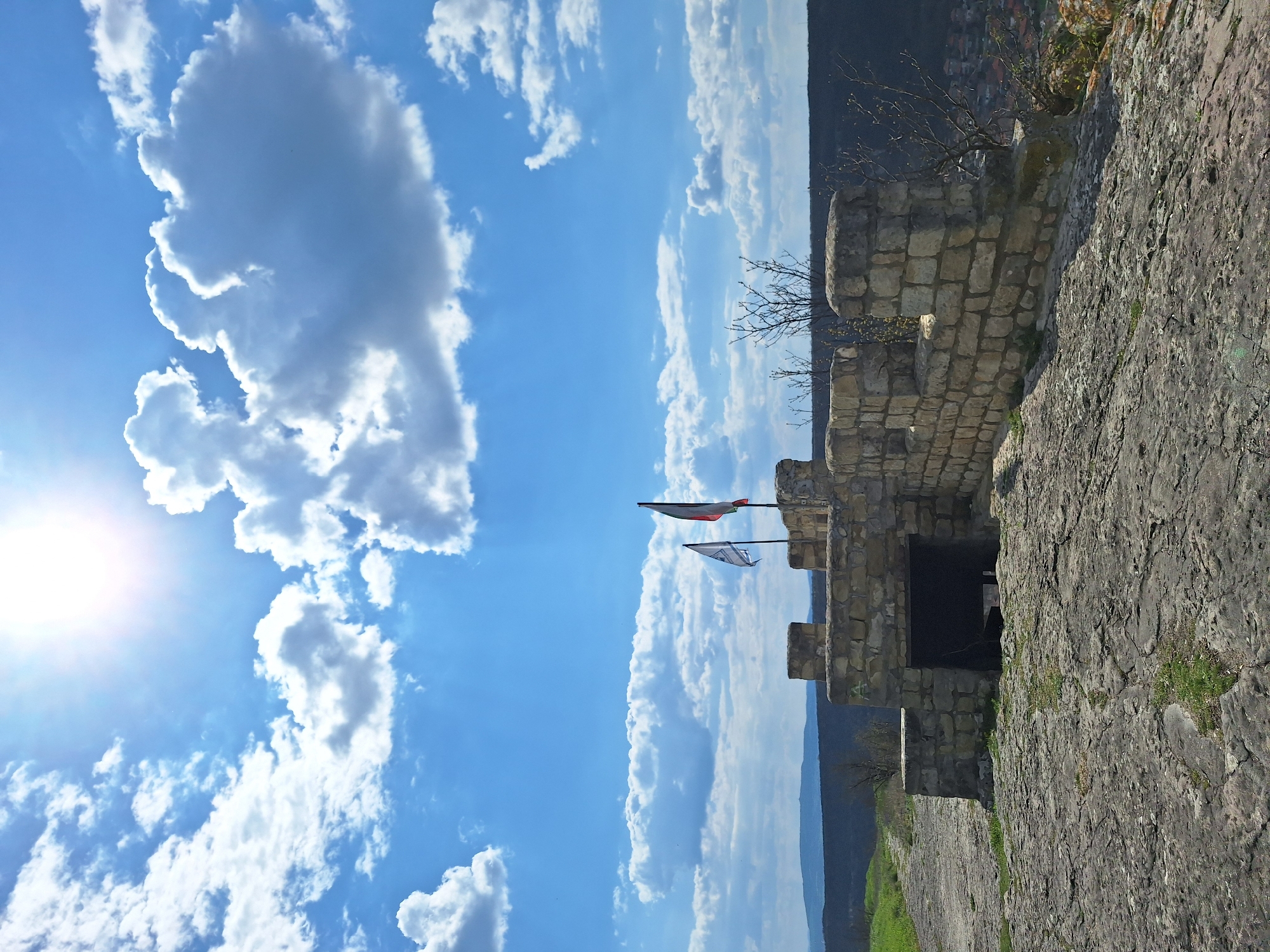 The image shows the Ovech Fortress in Provadia, Bulgaria, seen from a low angle, looking upward. The fortress wall, constructed from light-colored stone, runs along the right side of the frame. Two flags fly from the wall, against a backdrop of a bright blue sky with white clouds. The sun is visible in the upper-left corner. In the background, there is a view of the terrain outside the fortress walls.