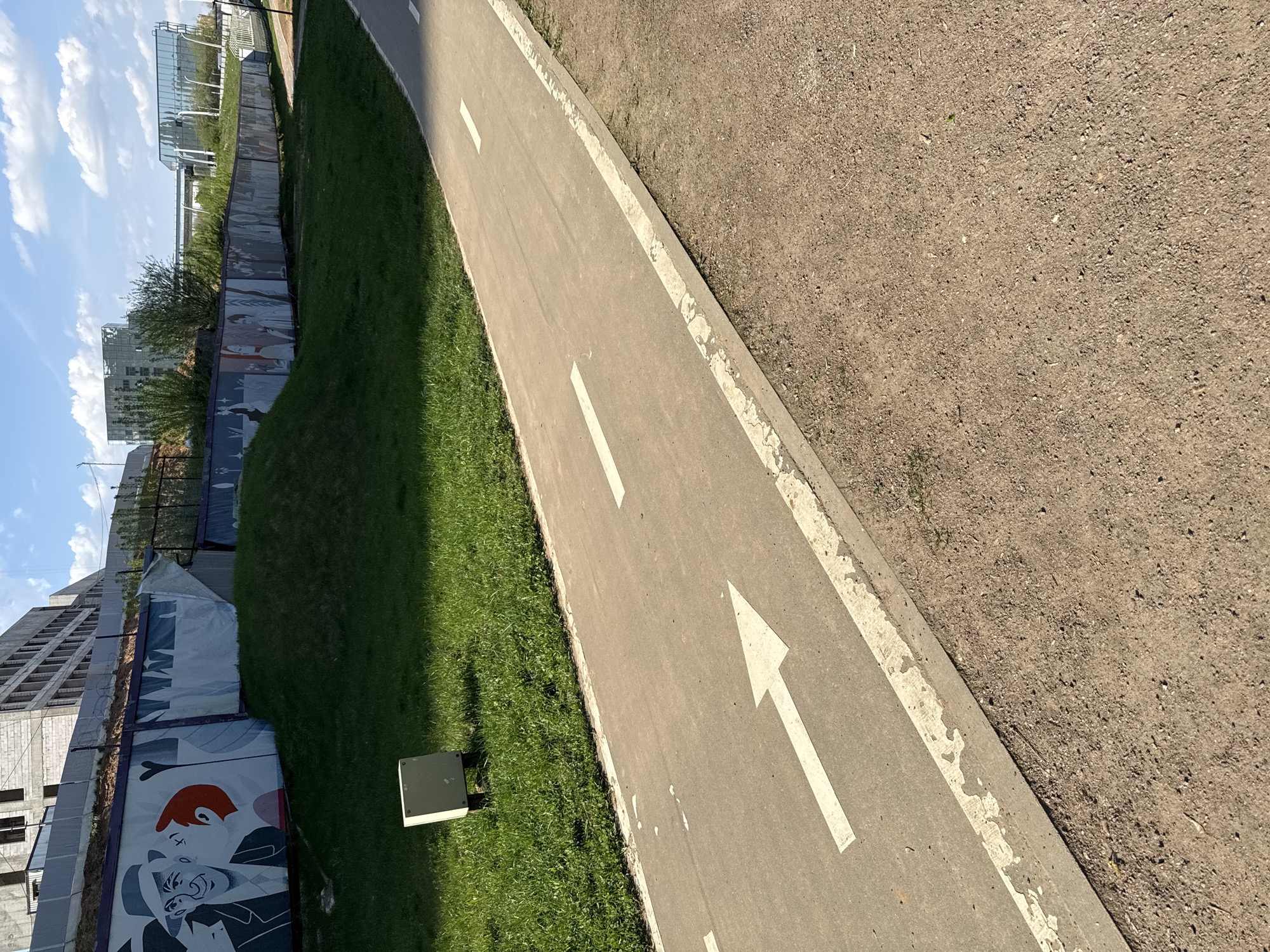 The image shows a section of a pedestrian and cycle path in Moscow, Russia. The path is made of asphalt and has white directional arrows painted on it. To the left of the path is a strip of grass and a concrete base for what appears to be electrical equipment. Further to the left is a temporary fence with graphic murals on it. Beyond the fence, construction work is visible on a large building. To the right of the path is an area of dirt. In the background, other buildings and sky are visible.