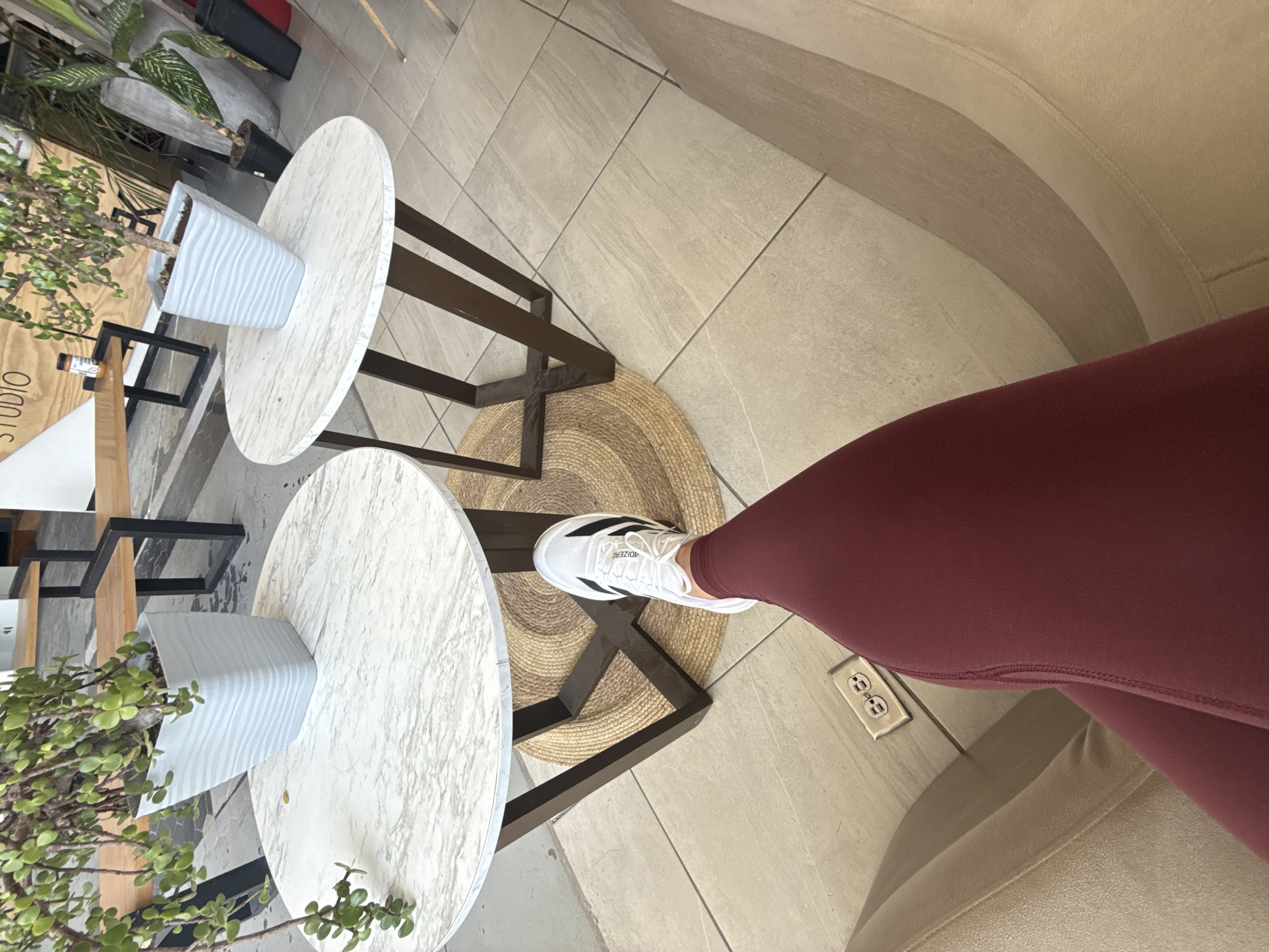The image displays a low-angle perspective from a seated position, primarily featuring a person's extended left leg and the immediate environment of a contemporary commercial space in Puebla, Mexico. The leg is clad in maroon athletic leggings and a white athletic shoe with three black stripes and "ADIZERO" branding.
Two identical small, round tables with white and gray marble-patterned tops and dark brown angular metal bases are prominent in the midground. Beneath the table closest to the viewer is a circular woven mat of natural fibers. Each table supports a light gray, square planter with a textured, rippled surface, containing a leafy green succulent-type plant. The plant on the left table exhibits significant downward growth.
The floor is tiled with large, light-colored square tiles, likely ceramic or porcelain, separated by dark grout lines. A metallic floor-mounted electrical outlet with two receptacles is visible on the floor in the lower left. The individual is seated in a light beige upholstered chair or bench, parts of which are visible in the lower corners.
In the background, a modern interior or semi-outdoor area is visible, featuring a wooden bench with a black metal frame. A white triangular sign with the partial word "STUDIO" in black lettering is mounted on a wooden panel behind the tables. Other potted plants in dark containers are also present in the background. The scene suggests a cafe, lounge, or common area within a commercial establishment or office building. The specific address within Puebla, Mexico, is not discernible from the visual information provided.