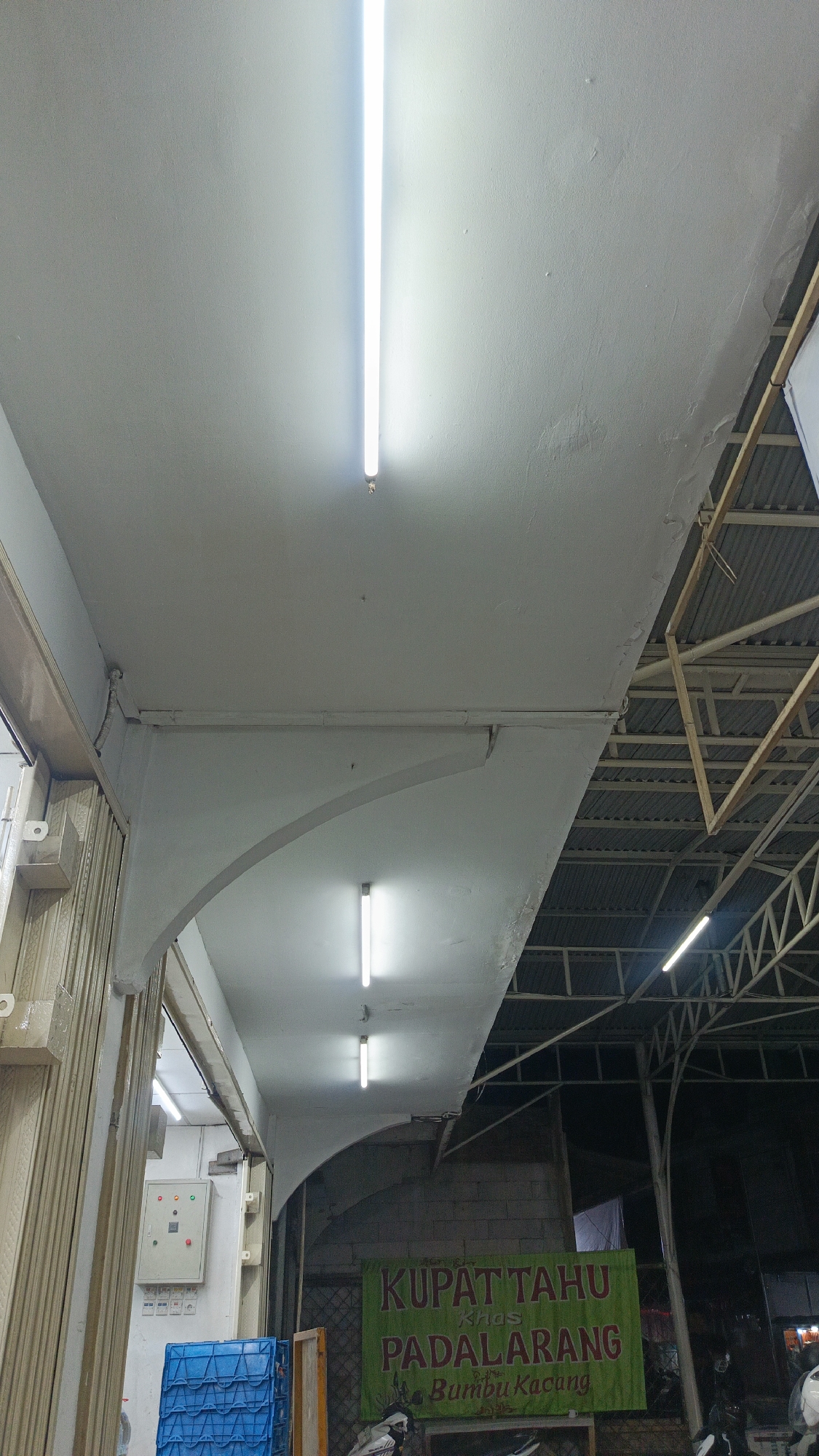 The image depicts an indoor marketplace setting in Margajaya, Indonesia. The scene is illuminated by fluorescent tube lights affixed to a white, textured ceiling. In the foreground, a corrugated metal shutter, painted a light beige color, is partially visible on the left side. Adjacent to it, a white electrical panel with colored buttons and indicators is mounted on a white wall. Below the electrical panel, a blue plastic crate is present.
A wide, green banner with red and yellow text hangs in the midground, displaying the words "KUPAT TAHU KHAS PADALARANG Bumbu Kacang." The banner is positioned against a brick wall. To the right of the banner, a dark, corrugated metal roof supported by a wooden and metal frame structure is visible, suggesting the marketplace is covered.
The overall environment appears to be a commercial space, likely a food stall or a small shop within a larger market. The lighting is functional, and the presence of the shutter and the electrical panel indicates a typical Indonesian market stall. The image is taken from a low angle, looking upwards towards the ceiling and banner. No people are clearly identifiable in the frame.