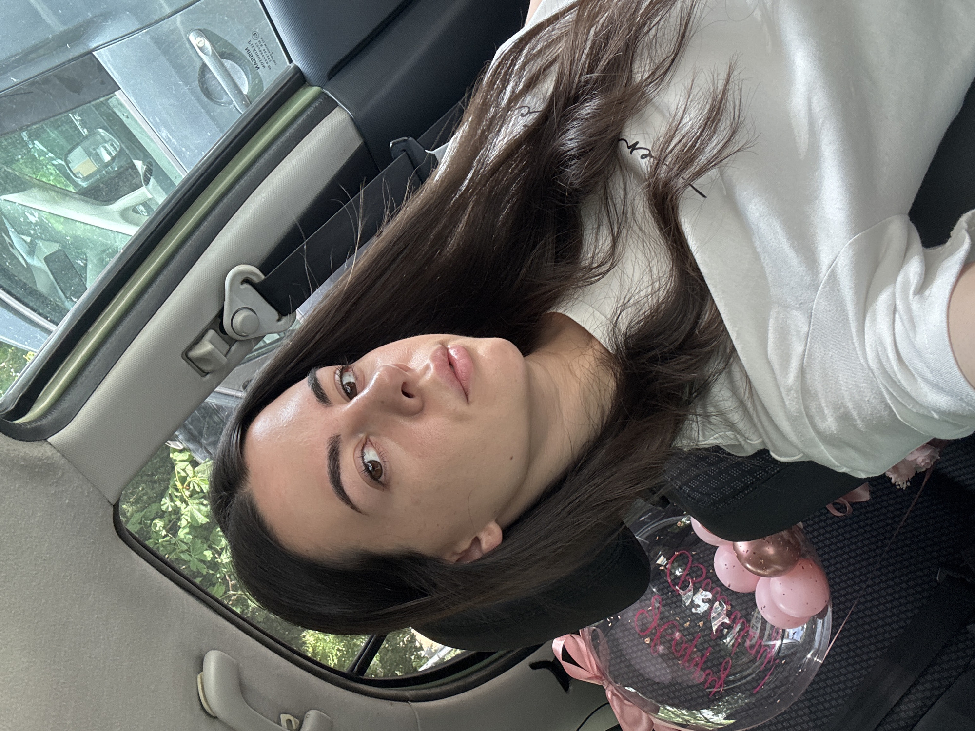 A medium-shot, slightly overhead selfie captures a young adult female with long dark brown hair inside a vehicle. She has light skin, dark eyebrows, and brown eyes, looking directly at the camera with a neutral expression and slightly parted lips. Her hair is parted in the middle and flows over her shoulders and chest. She is wearing a white short-sleeved t-shirt with dark text visible on the chest ("en" and part of another letter).
She is seated in the front passenger seat of a car, evidenced by the visible passenger-side door frame, window, and grab handle on the ceiling. The car's interior features a light grey fabric headliner and A-pillar, with a dark grey or black dashboard/door panel. A grey seatbelt mechanism is visible on the B-pillar.
Through the windows, lush green foliage from trees is visible, suggesting an outdoor, possibly urban or suburban, setting. A partial reflection of another vehicle, possibly a dark-colored SUV or sedan, is seen in the car's exterior, specifically showing a door handle and part of the side mirror. Text is faintly visible in reverse on the other vehicle's window: "NISSAN" and some smaller text below it.
Behind the woman's seat, on the rear seat or floor, a transparent bubble balloon is visible. Inside the balloon are several small pink and one rose-gold colored balloons, along with pink confetti. Pink script text, partially visible, reads "Happy Birthday". A pink ribbon is tied to the balloon's base, and a light pink object, possibly flowers, is also present near the balloon.
The scene takes place in a car in the city of Unknown, Unknown.