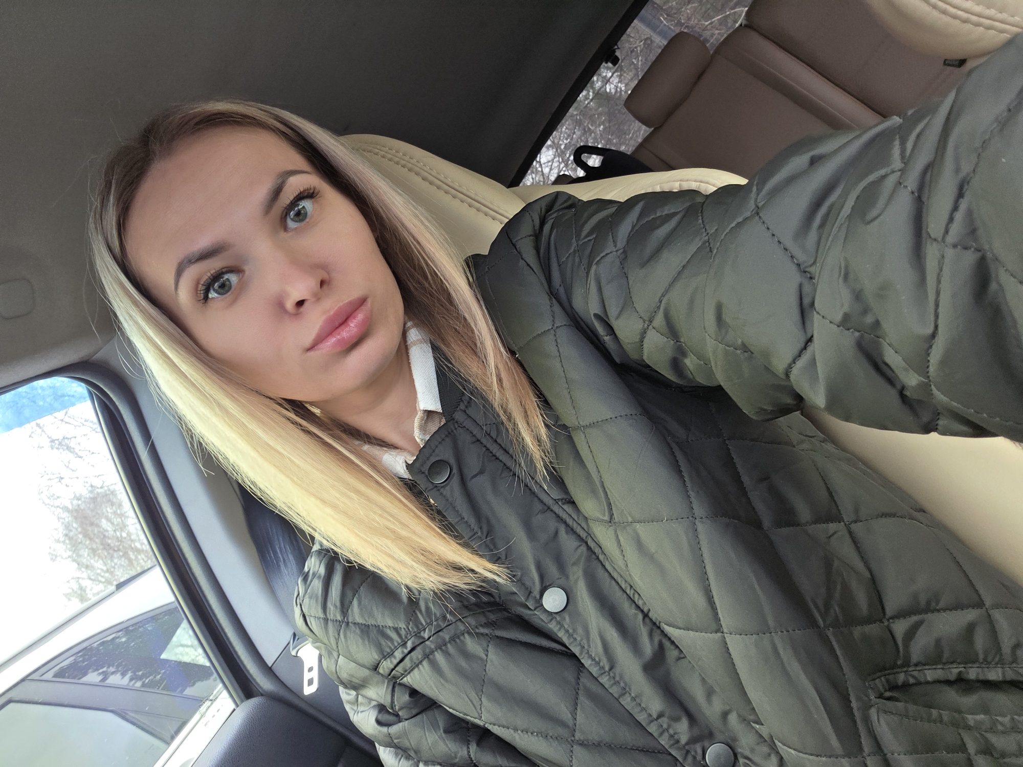 A woman is taking a selfie inside a car. She has blonde hair, light eyes, and full lips. She is wearing a black quilted jacket over a white shirt. The car interior is beige, and outside the window, there are trees and a building partially visible. There's no explicit indication of being in Chaikovsky, Russia.