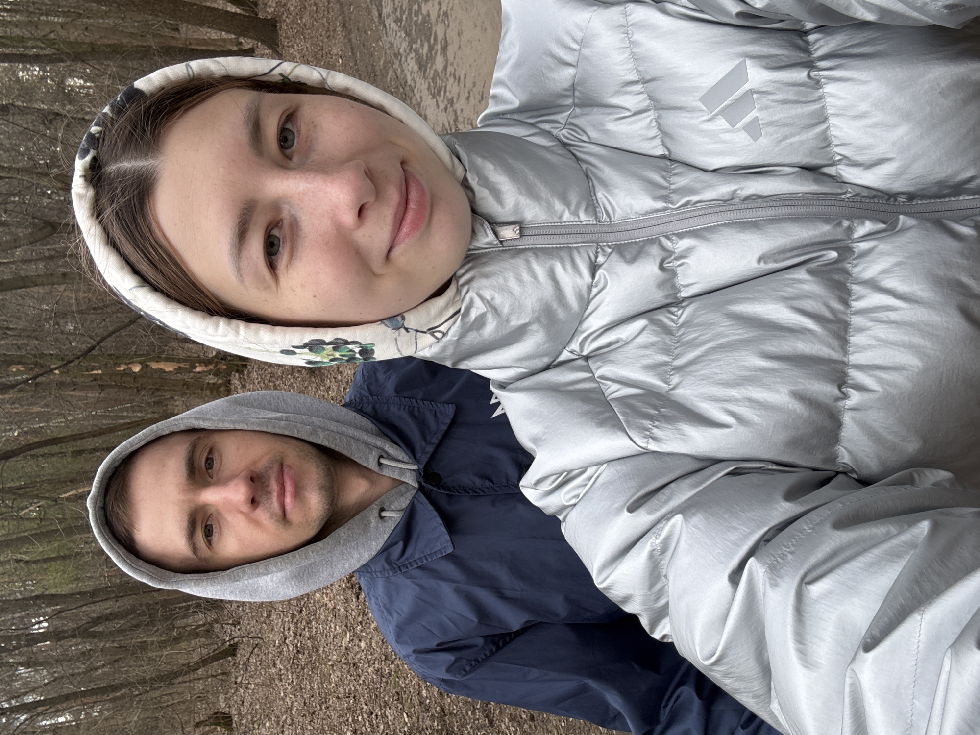 The image depicts two people outdoors, likely in a park or wooded area. A woman with light skin is wearing a shiny, silver puffer jacket with the hood up. A man with light skin is wearing a blue jacket with a gray hood. The background shows trees without leaves and a ground surface of dirt and dead leaves. The location is possibly in Moscow, Russia.