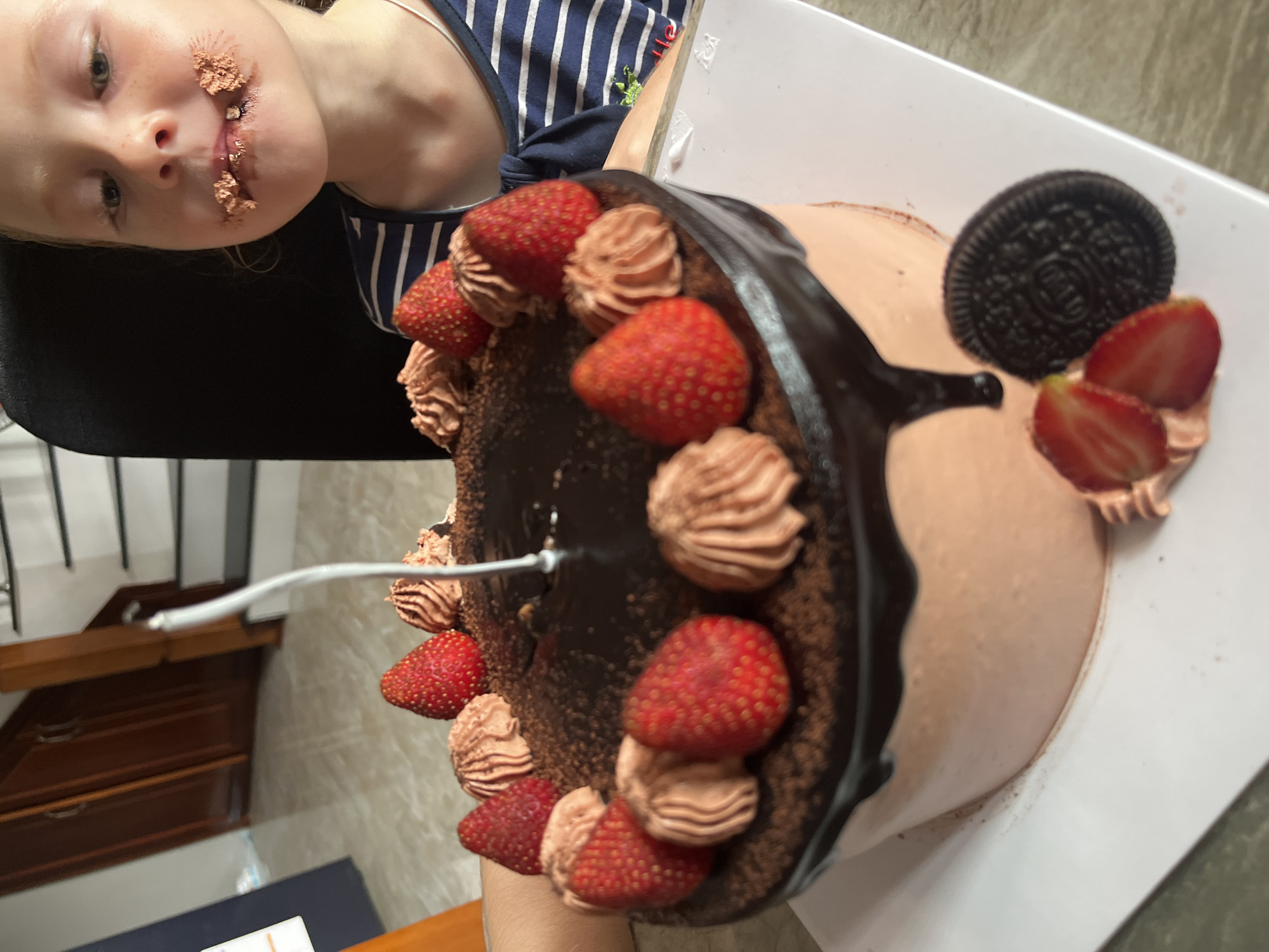 A child with chocolate smeared around the mouth is next to a chocolate cake decorated with strawberries, chocolate frosting swirls, and an Oreo cookie. The cake is on a white plate or surface. A metal object, possibly a fork or spoon, is near the cake. The location is Bac Nha Trang Ward, Vietnam. A wooden dresser is visible in the background, along with a staircase. The child appears to have been eating the cake.