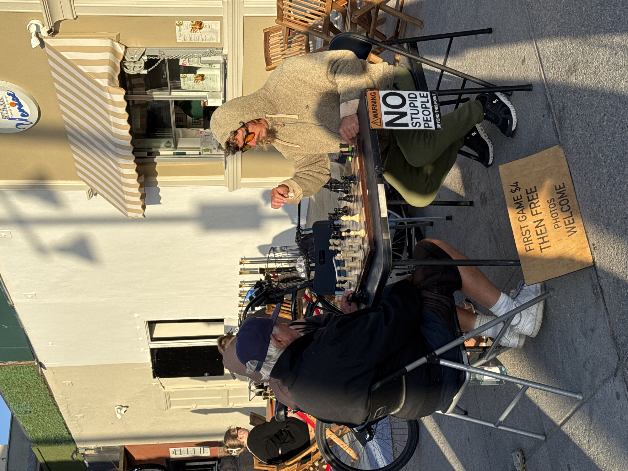 The image captures an outdoor scene on a sunny day, likely in Venice, Los Angeles, USA, depicting two men engaged in a game of chess on a sidewalk. One man wears a hooded sweatshirt and sunglasses, while the other dons a baseball cap. A sign propped against one of the chairs reads "WARNING NO STUPID PEOPLE BEYOND THIS POINT." Another sign on the ground indicates "FIRST GAME $4 THEN FREE PHOTOS WELCOME." A bicycle is situated near the chess game. In the background, a storefront with an awning labeled "STARA Pierogi & Sausage Venice" is visible. The setting is urban, and the scene appears to be a candid moment between the two chess players and the surrounding environment.
