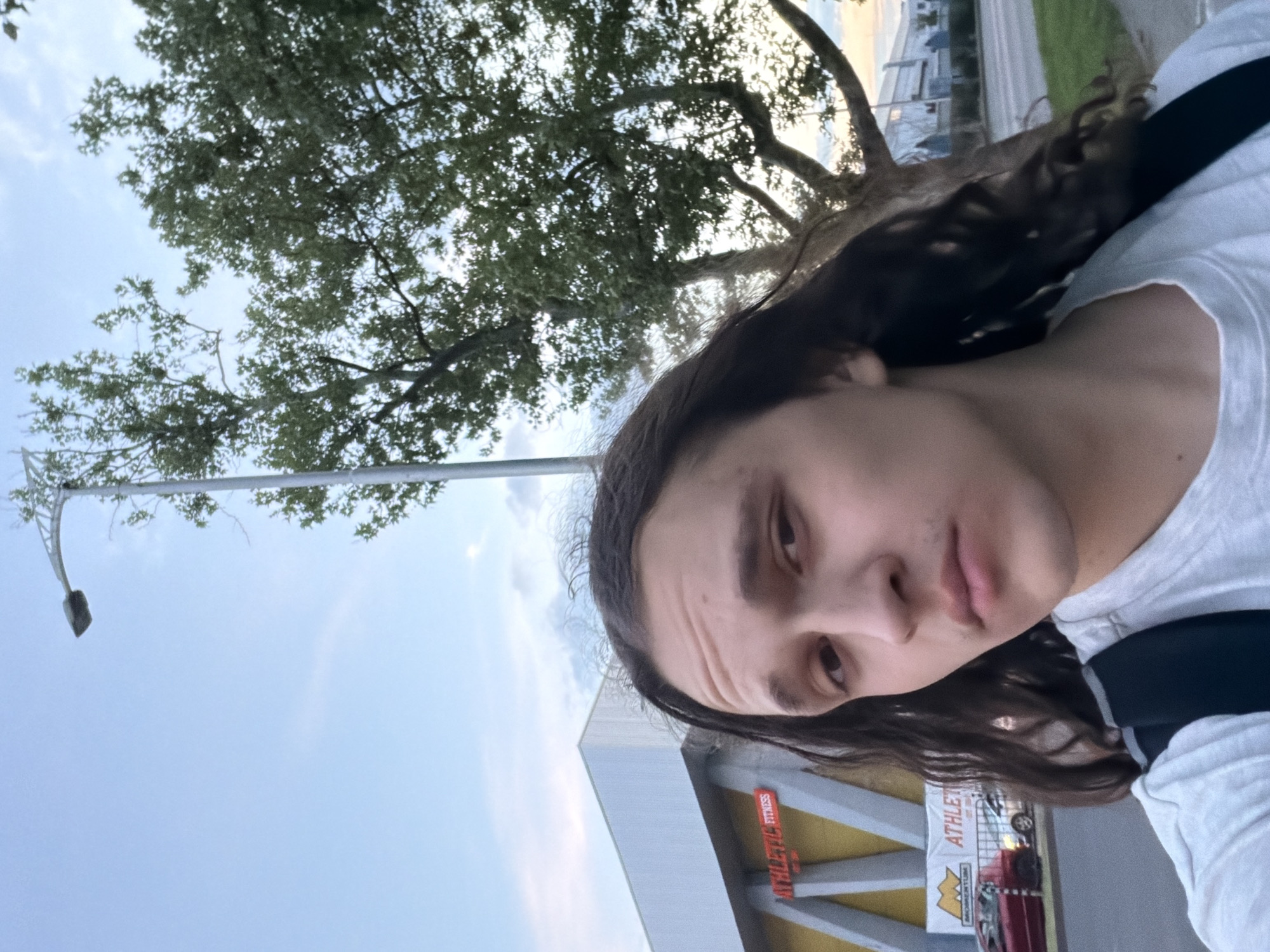 A selfie shows a person with long brown hair, wearing a white t-shirt with a black strap. The background includes a tree with green leaves, a light blue sky, a street light, a building with the sign "ATHLETIC FITNESS" and a few cars. The building is orange and grey. The photograph was likely taken in Plovdiv, Bulgaria based on the signage and overall style.
