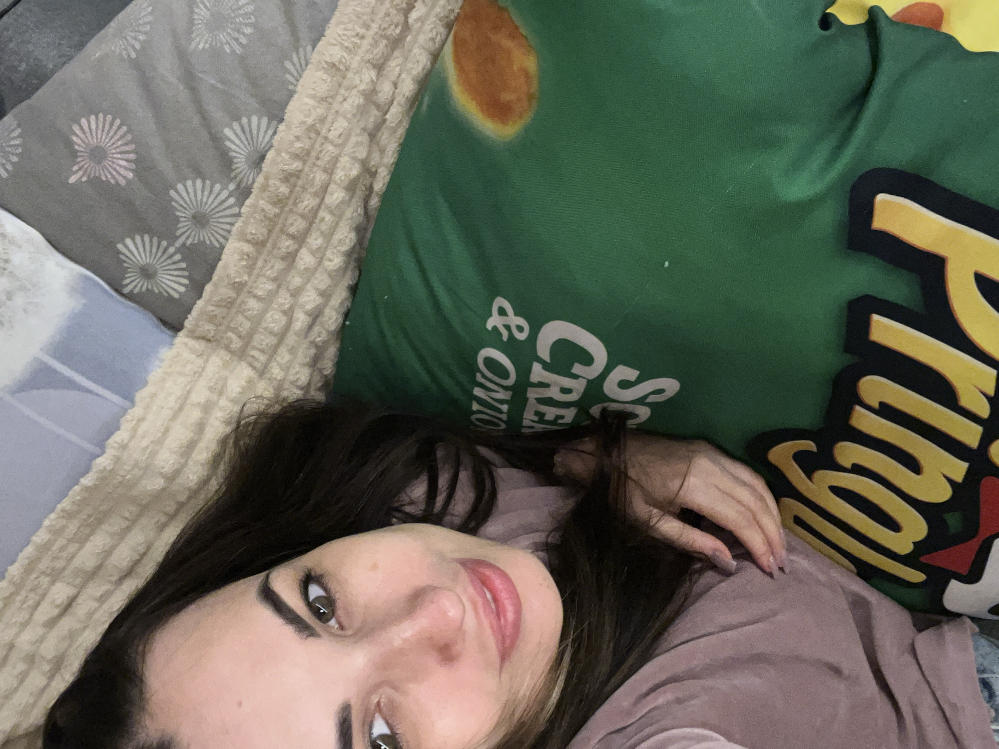 A close-up overhead shot shows a woman lying on her side on a bed. She has dark hair and is wearing a light pink t-shirt. Her eyes are open and she is looking at the camera with a slight smile. She is lying on a green pillow with white text and graphics. The text on the pillow appears to be "SCREAM & ONION" and a yellow and red graphic of a Pringles can. The background of the image consists of beige and grey patterned bedding. The scene appears to be indoors, likely in a bedroom.