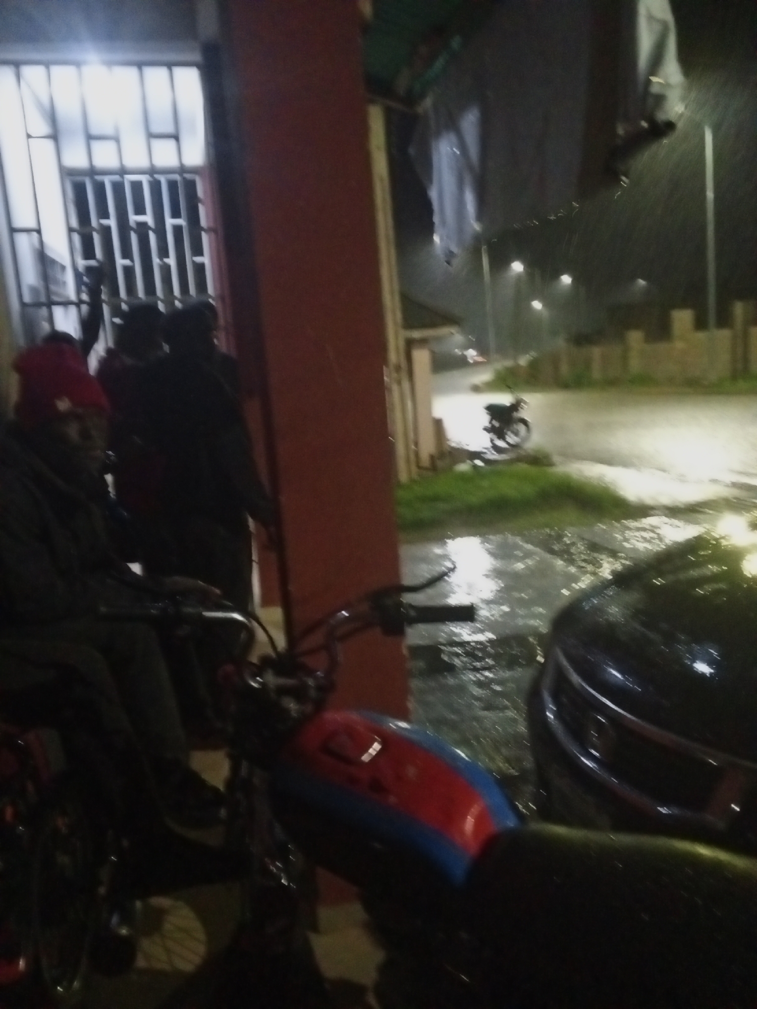 The image captures a scene at night in Gura Top, Nigeria, under heavy rainfall. In the foreground, the front of a motorcycle with a red, blue, and black fuel tank is visible. To the left, a person wearing a red beanie and dark clothing is seated on a bicycle. Behind them, partially obscured by a red pillar, are several other individuals standing and appearing to converse. To the right, the front of a dark-colored car is parked, its surface glistening with rainwater. The background depicts a street with puddles and reflections of streetlights. A motorcycle is parked on the side of the street. Clothes are seen hanging from an overhead line. The overall environment suggests a residential or commercial area experiencing inclement weather.
