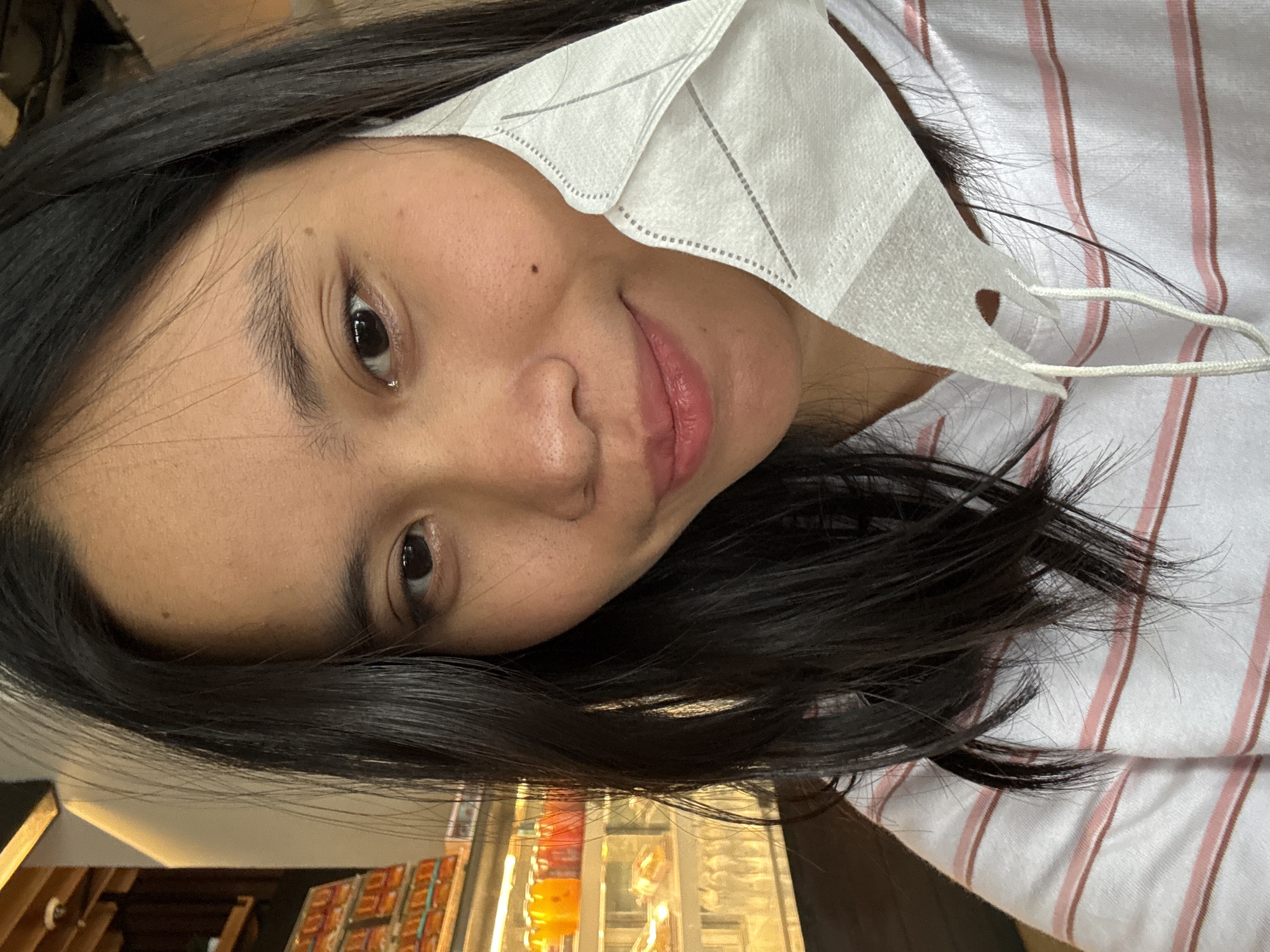 A young woman is taking a selfie indoors, with a blurred background that appears to be a cafe or restaurant. She has dark hair and is wearing a white hoodie with thin pink and brown stripes. She is holding a white face mask partially covering the right side of her face. The environment suggests a public indoor space, possibly in East Jakarta, Indonesia, given the context.