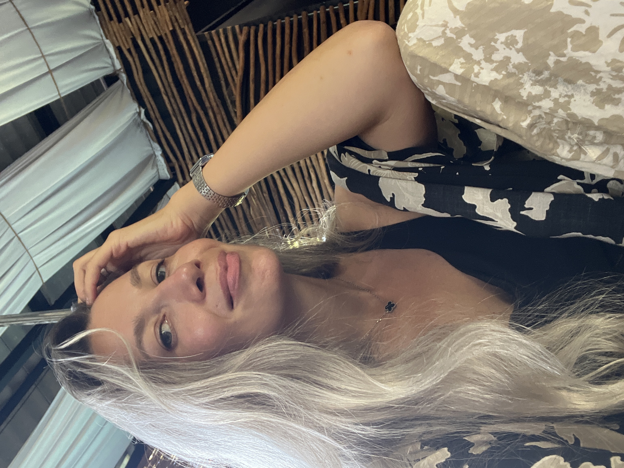 This is a close-up, portrait-oriented photograph of a young woman with long, blonde hair. She is lying down, with her head tilted and one hand resting on her forehead. Her eyes are open and she is looking towards the camera. Her lips are slightly parted, and she appears to be in a relaxed or playful mood. She is wearing a black top with a white pattern and a silver watch on her left wrist. A delicate necklace with a clover pendant is visible on her collarbone.
The background is out of focus but suggests an indoor setting. To the left, there are white fabric drapes, possibly part of a tent or canopy. To the right and behind her, there are vertical wooden elements, resembling reeds or bamboo, and a light-colored patterned fabric, possibly a cushion or upholstery.
The lighting suggests it could be daytime, but it is difficult to determine the exact time of day or weather conditions. The scene does not offer strong location cues beyond the potential for a resort or stylized interior, making the provided context of Weligama, Sri Lanka, the primary identifier of location. No visible text is present in the image.