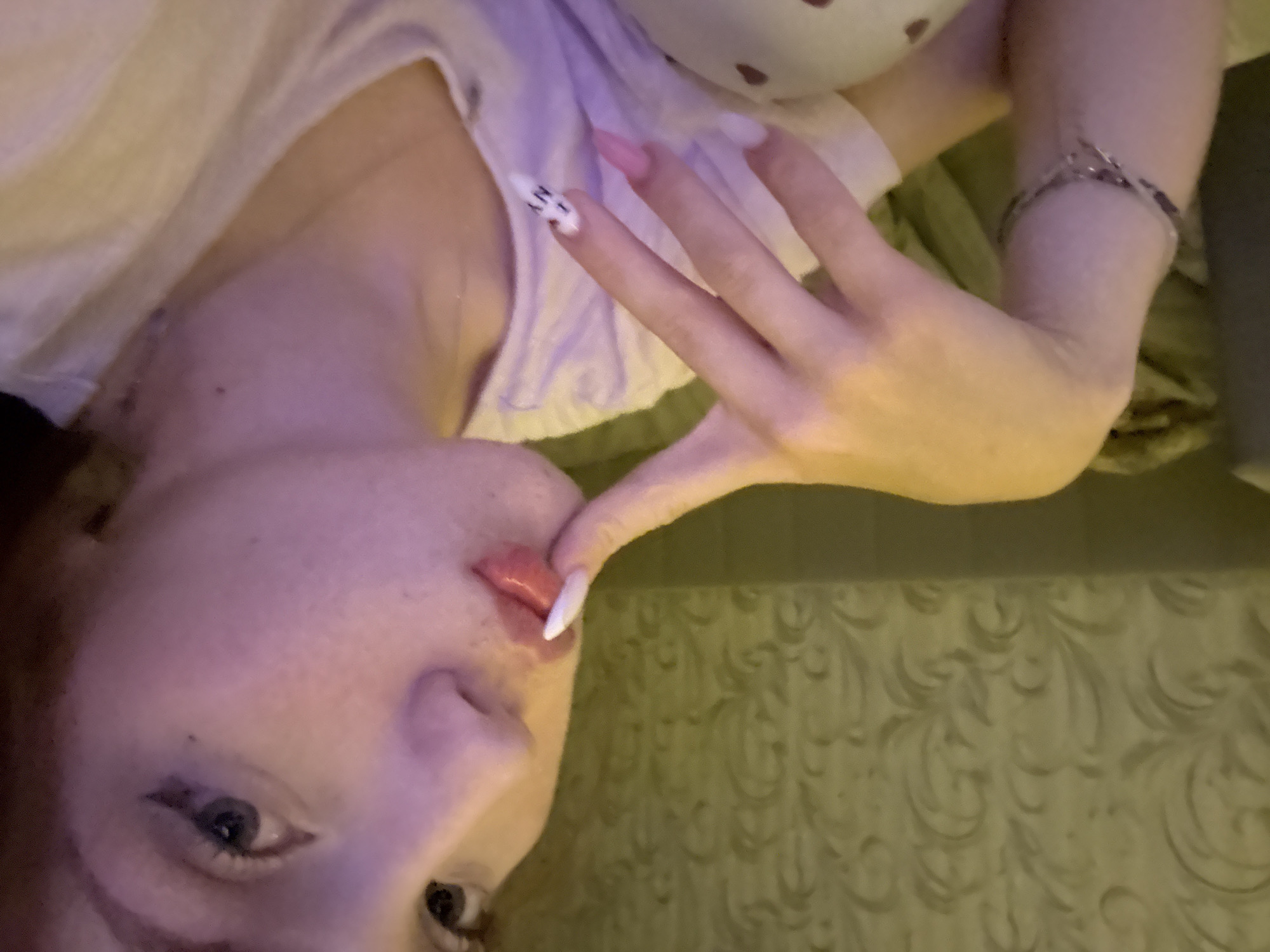 An indoor, close-up, eye-level selfie taken in Naberezhnye Chelny, Russia, features a young female subject. The subject has fair skin with visible freckles, light blue or grey eyes, and reddish-brown hair partially visible on the right. She wears black winged eyeliner and a small mole is visible below her left eye. Her full lips are lightly colored pink/red. She poses with her left index finger, adorned with a white, long, pointed nail, pressed against her lower lip. Other nails on her left hand are pink, with the pinky finger featuring a white base with black text or graphic art. A silver-toned bracelet with small decorative elements is on her left wrist. She wears a light-colored, possibly white or pale pink, crew-neck top, with a thin necklace chain faintly visible. The background consists of light olive green or muted gold wallpaper with a subtle, embossed, classical-style swirling pattern. Below the wallpaper is a horizontal section of darker, plain material. Warm artificial lighting illuminates the scene.