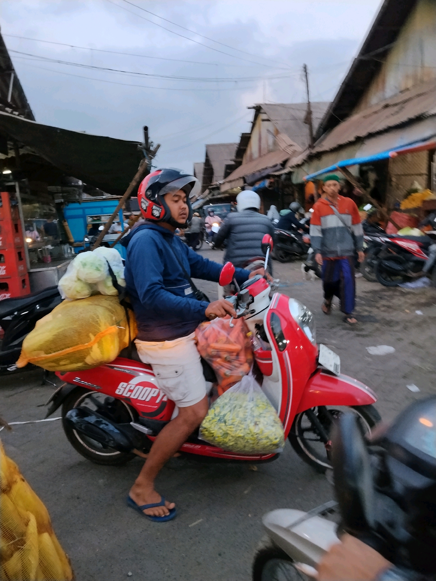 In the city of Malang, Indonesia, a man wearing a blue long-sleeved shirt, white shorts, and a red helmet rides a red scooter. The scooter is laden with produce, including a large yellow net bag filled with what appears to be gourds or squash, and a clear plastic bag containing green vegetables, possibly cabbage or lettuce. Another bag of carrots is visible on the right side of the scooter. The man is in the process of navigating a market area, with stalls and other people on scooters visible in the background. The sky is overcast.