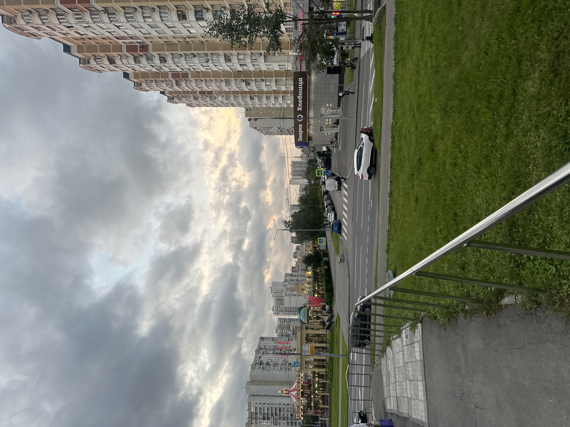 The image depicts a street scene in Moscow, Russia, during a cloudy day with the sun attempting to break through the overcast sky. The perspective is from an elevated position, looking down onto a street lined with tall, multi-story residential buildings. These buildings have a uniform architectural style with repetitive balconies and windows.
A white car is parked on the side of the road, and several other vehicles are visible further down the street. A pedestrian crossing is marked on the road, with a few people walking. Adjacent to the road is a strip of green grass, and beyond that, more buildings can be seen in the distance.
A prominent feature is a sign with Russian text, which reads "Пекарня Хлебница" (Pekarnya Khlebnitsa), indicating a bakery. There's also a carousel visible in the foreground, suggesting a recreational area or park nearby. The overall atmosphere appears calm and urban, with the cloudy sky lending a somewhat muted mood to the scene. The time of day is likely late afternoon or early evening, given the soft light.