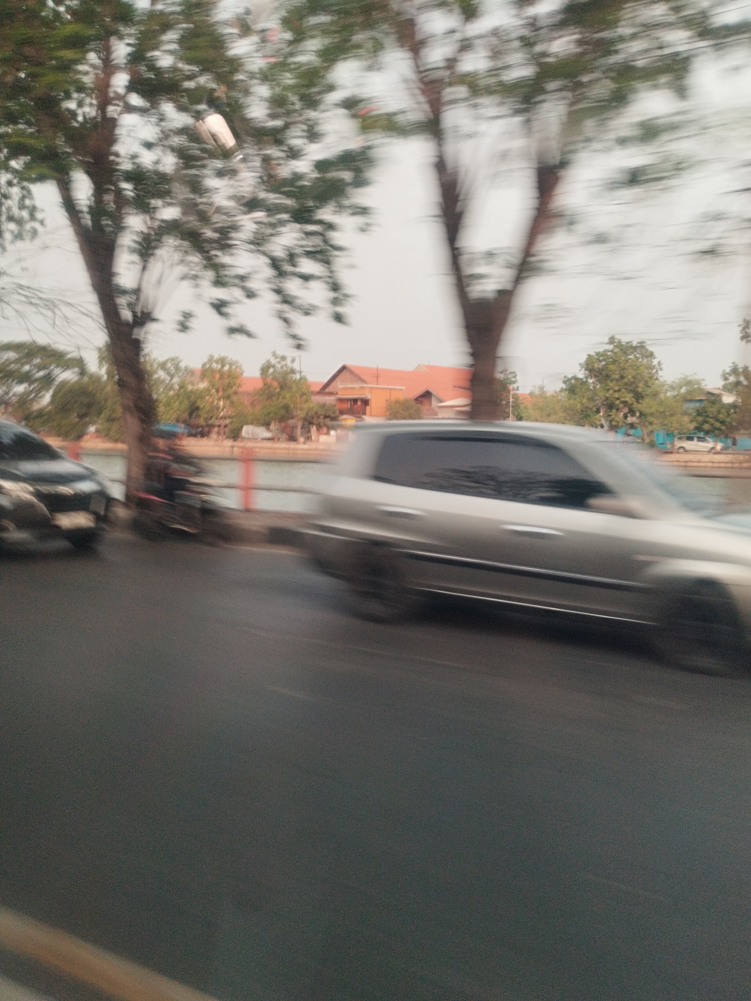 The image depicts a street scene with motion blur, suggesting movement. In the foreground, a dark-colored car is partially visible on the left. Further to the right, a silver car is more prominent, also showing significant motion blur. Behind the cars, a body of water is present, with a red railing along its edge. Trees line the opposite bank, with buildings, some with orange roofs, visible in the background. A motorcycle rider is also present, further blurred by motion. The overall environment appears to be an urban or suburban area in Surabaya, Indonesia, possibly near a canal or river.