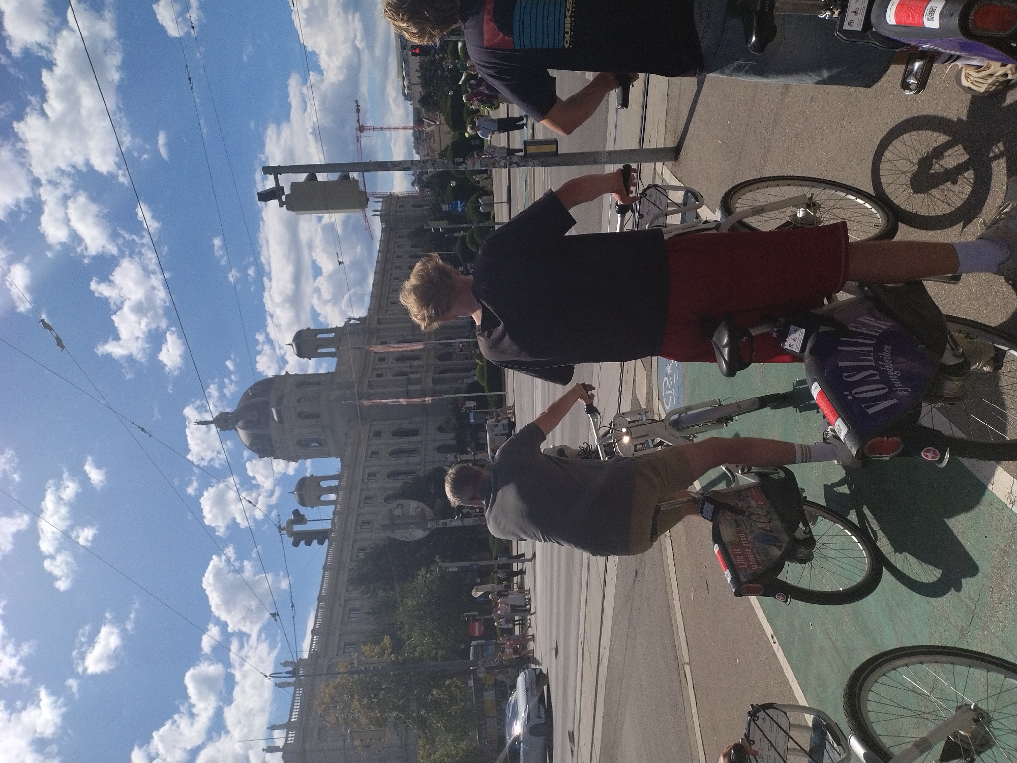The image captures a sunny day in Vienna, Austria, with a large, ornate building in the background, likely a museum or governmental structure. In the foreground, three individuals are on rented bicycles, navigating a street with tram tracks. The first person, seen from behind, is wearing a grey t-shirt and beige shorts, riding a white bicycle. The second person, also from behind, is wearing a dark t-shirt and red shorts, astride a white bicycle. The third person, whose back is mostly obscured, is wearing a dark t-shirt and denim shorts, also on a bicycle. The bicycles appear to be part of a shared rental system, as indicated by the signage on their rear racks. People and vehicles are visible in the distance, suggesting an active urban environment. The sky is blue with scattered white clouds, and overhead power lines crisscross the frame. The scene is a typical street view in a European city, focusing on urban mobility and sightseeing.