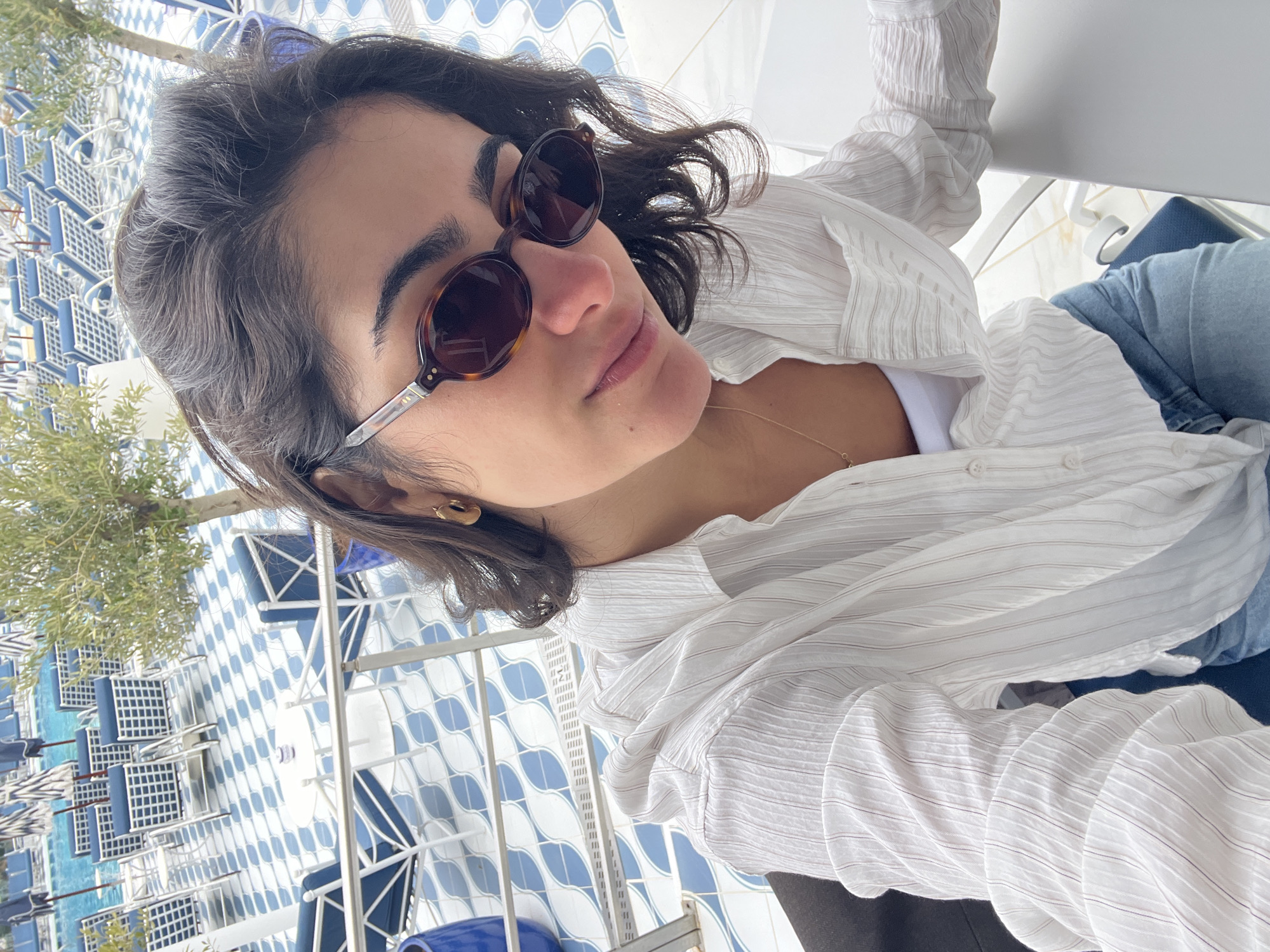 A woman with short, dark hair and sunglasses is looking up at the camera. She is wearing a white button-down shirt over a white tank top and blue jeans. She has a gold necklace and small hoop earrings. The background shows an outdoor patio with blue and white tiled tables and chairs, suggesting the image was taken at a restaurant or cafe. The location is Santa Eulària des Riu, Spain.