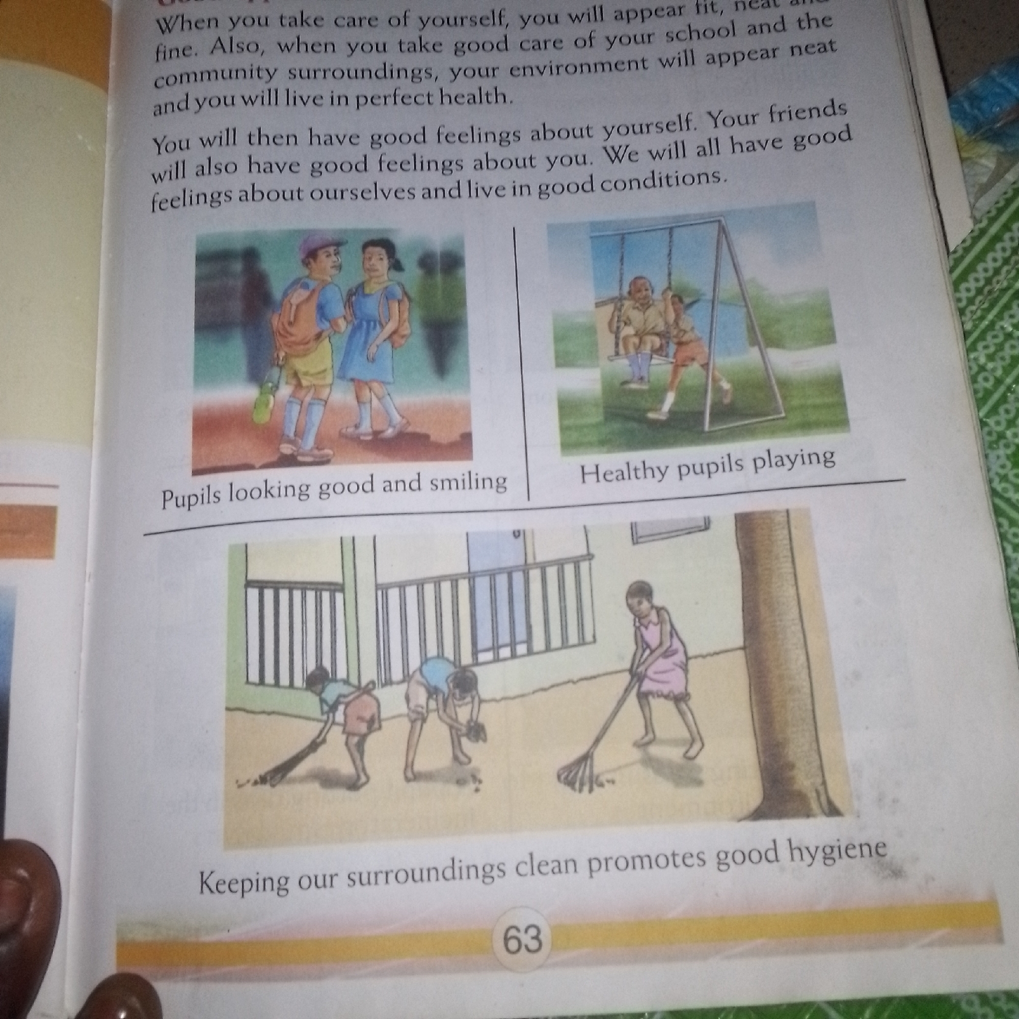 The image depicts a page from a children's book or educational material. The page contains text and three illustrations. The text discusses taking care of oneself and one's surroundings, and how this leads to good feelings and living in good conditions.
The top left illustration shows two children, a boy and a girl, walking with backpacks. The boy is wearing a yellow shirt and shorts, and the girl is wearing a blue dress. They appear to be on a sidewalk or paved area, with a blurred background that might suggest a school or park. Below this illustration is the caption "Pupils looking good and smiling".
The top right illustration shows two children on a swing set. One child is pushing the other. They are in an outdoor setting with grass and trees in the background. Below this illustration is the caption "Healthy pupils playing".
The bottom illustration depicts a group of children cleaning their surroundings. One child is raking leaves, while others are picking up debris from the ground. They are in an outdoor area with a building in the background that has a barred window. Below this illustration is the caption "Keeping our surroundings clean promotes good hygiene".
At the very bottom of the page, there is a circled number "63", indicating the page number. The overall impression is of educational content aimed at children, focusing on health, hygiene, and positive social interactions. The scene is not specific to any known city or location.