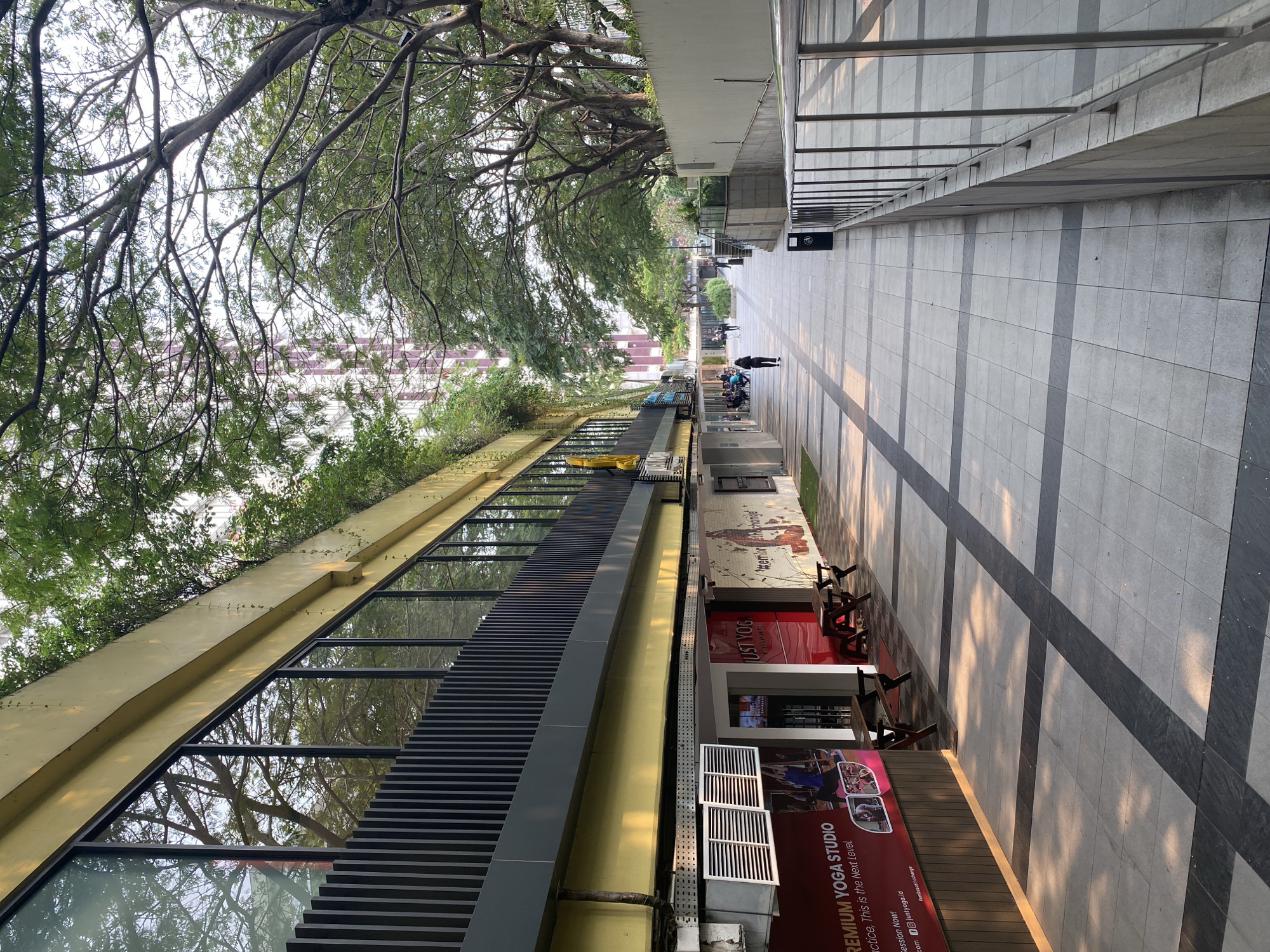 The image depicts a sun-dappled pedestrian walkway in front of a building with a yellow facade. Large windows reflect the surrounding trees and a tall, modern building in the background. Greenery cascades from the top of the building, creating a natural overhang. The walkway is paved with square tiles in a grid pattern, with alternating dark and light gray tiles. Benches and picnic tables are placed outside a yoga studio with prominent signage. Several people are visible walking along the path, some singly and others in small groups. A trash receptacle is positioned near a metal railing that lines the right side of the walkway, which appears to descend slightly. The overall atmosphere suggests a pleasant outdoor space within an urban setting. The yoga studio is identified as "PREMIUM YOGA STUDIO" and "JUST YOGA," with the tagline "practice, This Is the Next Level." The location is specified as North Jakarta, Indonesia.