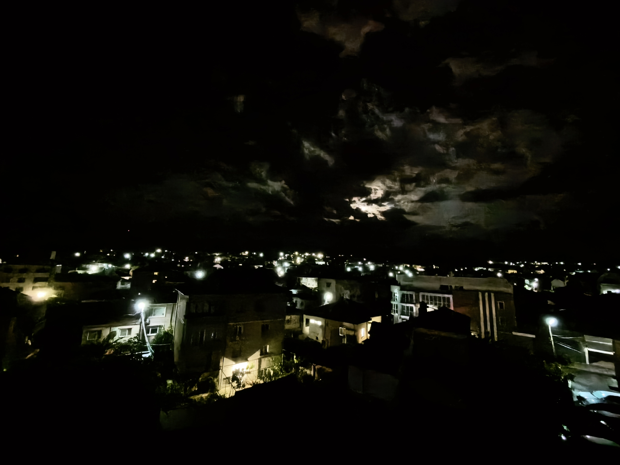 The image shows a night view of a city, possibly residential, under a cloudy sky where the moon is trying to shine through. Numerous streetlights and lights from buildings illuminate the scene. The rooftops of houses are visible, varying in height and shape, some with trees and vegetation nearby. Some cars are visible at the bottom-right corner. The city and specific location are unknown.