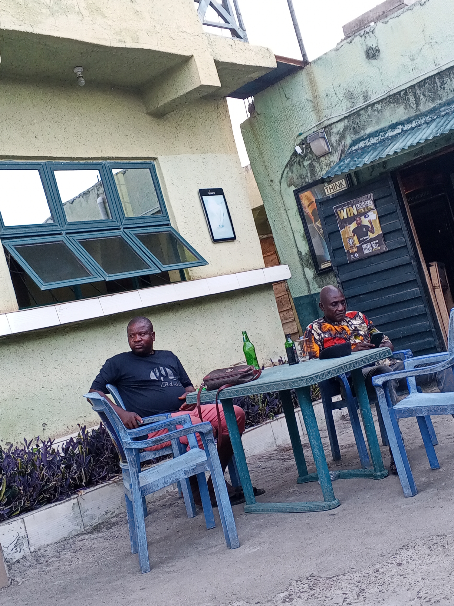 This image captures two men seated at a plastic table outdoors, likely at a casual bar or eatery in Zango-ndaji, Nigeria. The scene is set against two weathered buildings; one is light yellow/beige with large, open-vent windows, and the other is light green with a dark wooden doorway and a corrugated metal awning.
The man on the left wears a black T-shirt with "Animal" and "Adidas" visible text, and red shorts. He is seated on a blue plastic chair, looking away from the camera. Beside him on the green plastic table are two green bottles, one possibly a beer bottle, and a small dark bag.
The man on the right, bald, wears a colorful patterned shirt and dark trousers. He is also seated on a blue plastic chair, engrossed in his smartphone. A glass is on the table in front of him. Both men appear relaxed, possibly socializing or simply passing time.
The sky is overcast, suggesting daytime without direct sunlight. The ground is paved, and a small garden bed with purple-leafed plants borders the area.
Notable details include signs on the green building: a poster with text "THINK" and "WIN A CHILLING..." along with a "Guinness" logo and "STEPS TO STAND A CHANCE TO WIN". Another smaller sign or screen with "Guinness" is visible on the light yellow building. A security camera and an outdoor light fixture are also visible on the buildings. The plastic furniture, weathered buildings, and overall informal setting contribute to the local ambiance.