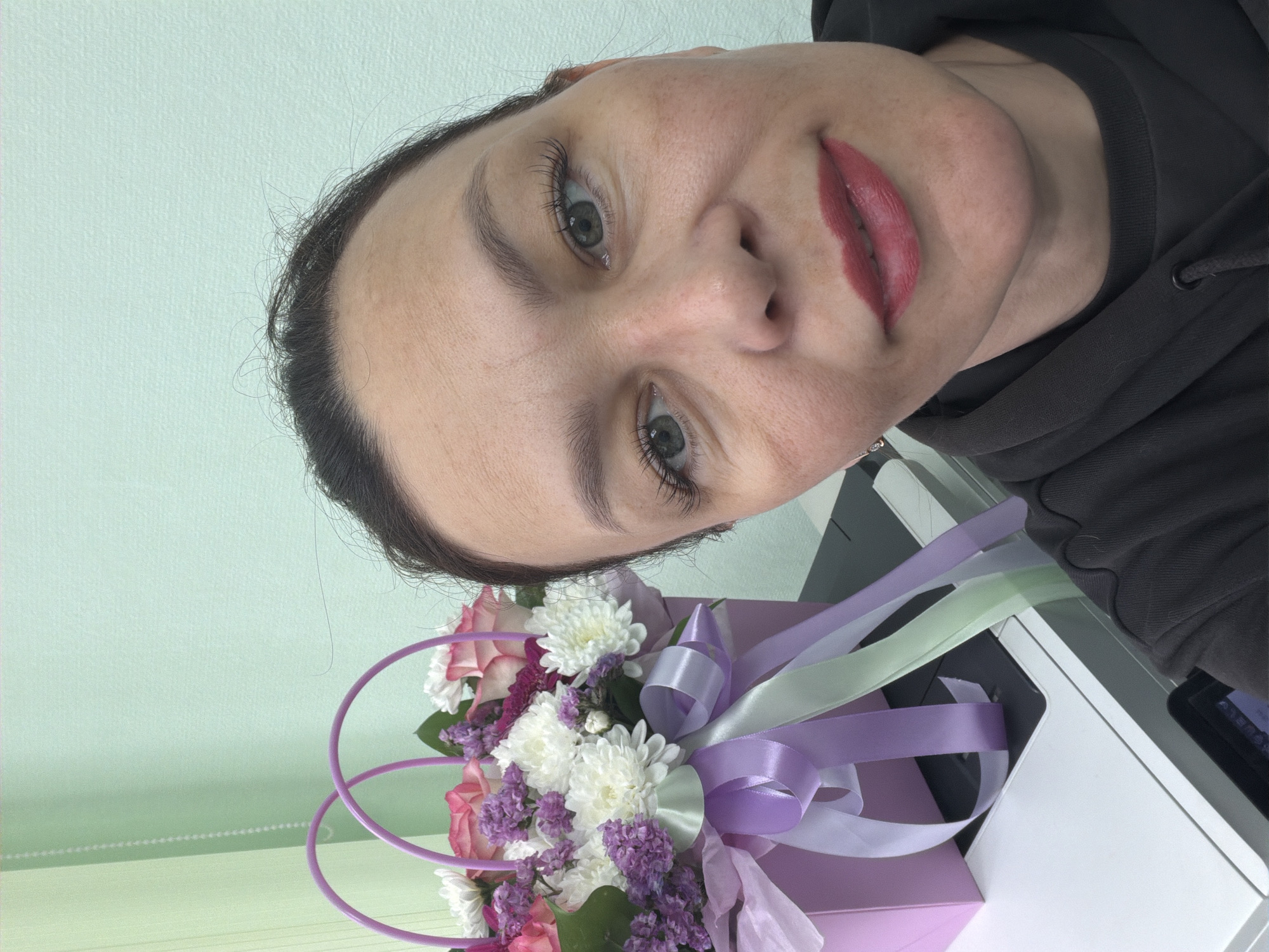 A woman poses with a bouquet of flowers. The bouquet contains roses, chrysanthemums, and other small purple flowers, arranged in a purple box with lilac, silver, and light green ribbons. The background is a pale green wall. The scene is located in the city of Chaikovsky, Russia.