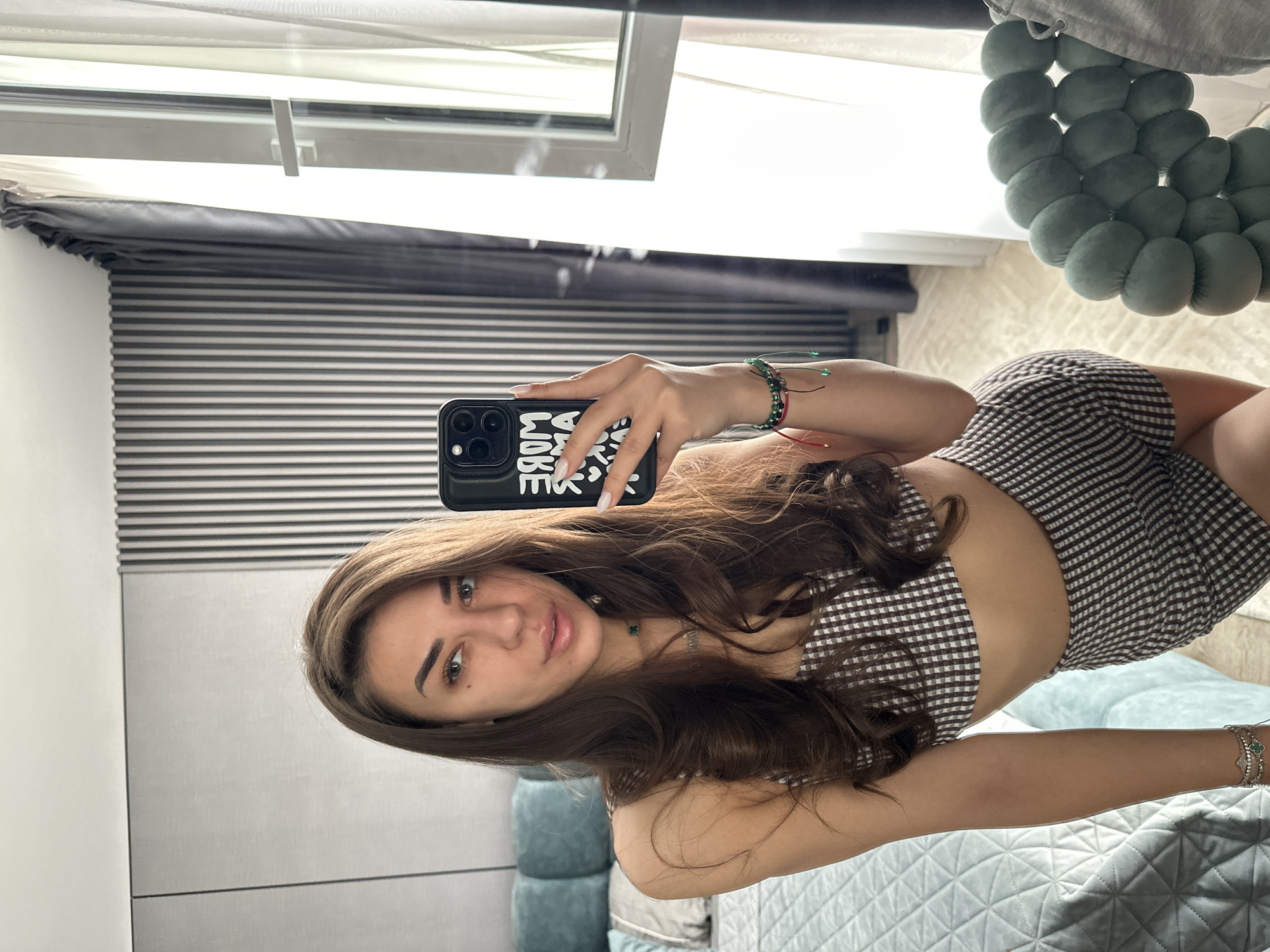 A young woman with long, dark brown hair, wearing a brown and white checkered crop top and matching shorts, is taking a selfie in a mirror. She is wearing bracelets on her left wrist and a necklace. In the background, a bed with a light blue quilted comforter and pillows is visible, along with a window with sheer white curtains and darker gray drapes. The room appears to be a bedroom, possibly in a modern apartment. The location is not specified within the image or context provided, thus cannot be identified as Sofia, Bulgaria.