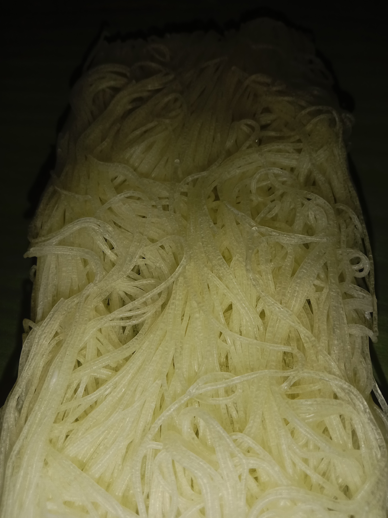 The image is a close-up, vertical shot of a pile of uncooked rice vermicelli noodles. The noodles are thin, white, and coiled in various directions, creating a textured surface. The lighting is dim, with shadows obscuring parts of the pile and the background, which appears to be a dark surface, possibly a table or countertop. The noodles have a slightly translucent quality and a ribbed texture along their length. There are no people or specific objects identifiable beyond the noodles themselves, and no context is provided regarding their location, including the city of Lemahkembar, Indonesia.