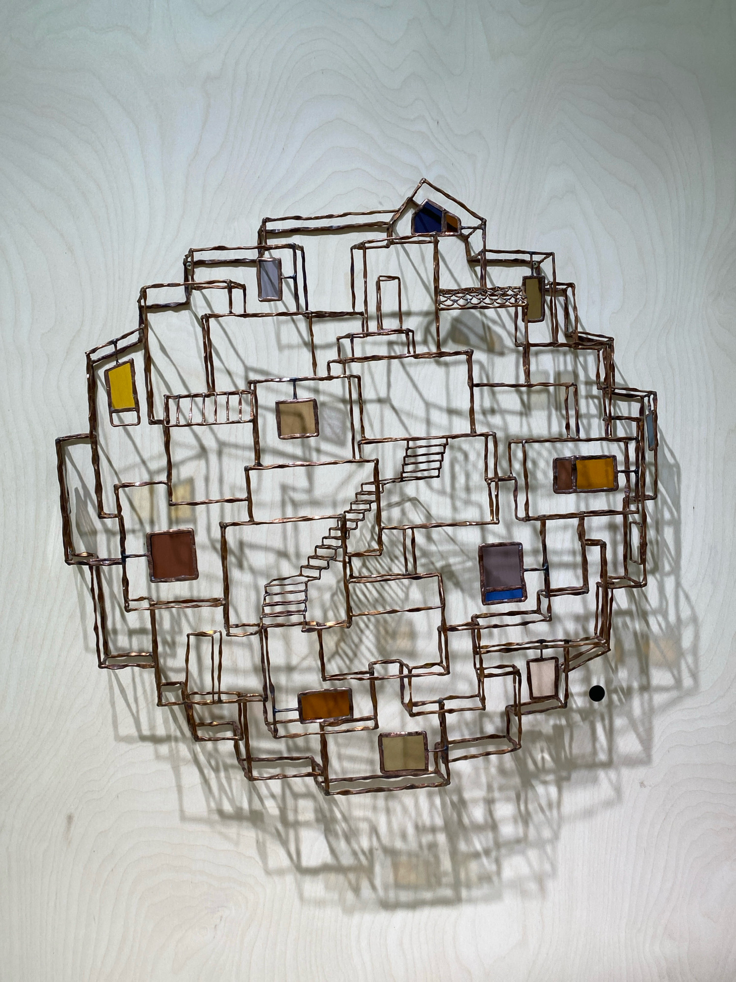 The image shows a piece of copper-framed stained glass art mounted on a light-colored wooden wall. The artwork is abstract, resembling a cluster of interconnected structures with various colored panes of glass representing windows or openings. The shapes are geometric, forming a somewhat irregular, circular outline. The glass panes are in shades of yellow, brown, blue, and clear or lightly colored. Shadows from the artwork are cast onto the wooden wall, creating a dynamic visual effect. The background wall has a visible wood grain pattern. There are no people or other identifiable objects in the scene besides the artwork and the wall. There is no information within the image that suggests a location in Khimki, Russia.