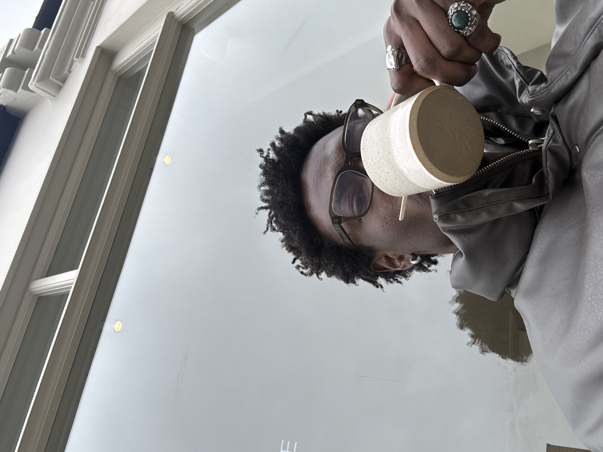 The image shows a person with dark curly hair, wearing glasses and a gray leather jacket, holding a speckled beige mug with writing on it. The person has multiple rings on their hand. The scene appears to be viewed through a glass window, which reflects a blurred interior and the person holding the mug. On the left side of the window is a section of a building facade. There is no indication in the image that the location is in London, United Kingdom.