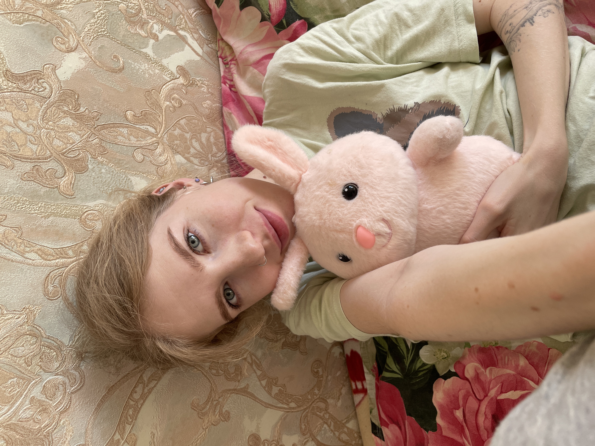 An overhead shot shows a young individual lying on a bed or sofa, looking directly at the camera with a gentle expression and a slight smile. The person has light blonde hair, striking blue eyes, and fair skin. They wear a light green t-shirt with a dark, stylized graphic design on the chest. Visible on their left arm is a fine-line tattoo, and their left ear features multiple piercings including helix, tragus, and lobe, along with a septum piercing on their nose.
They are holding a plush pink bunny toy, which has black bead eyes and a soft pink nose, cuddled against their chest. The background consists of two distinct patterns: the surface they are lying on is a light gold and beige patterned fabric with intricate damask or floral motifs, while a blanket or duvet with large, vibrant pink and red floral patterns on a dark background is partially visible behind them and to their right. The lighting is soft and even, suggesting an indoor setting. No text is visible in the image.