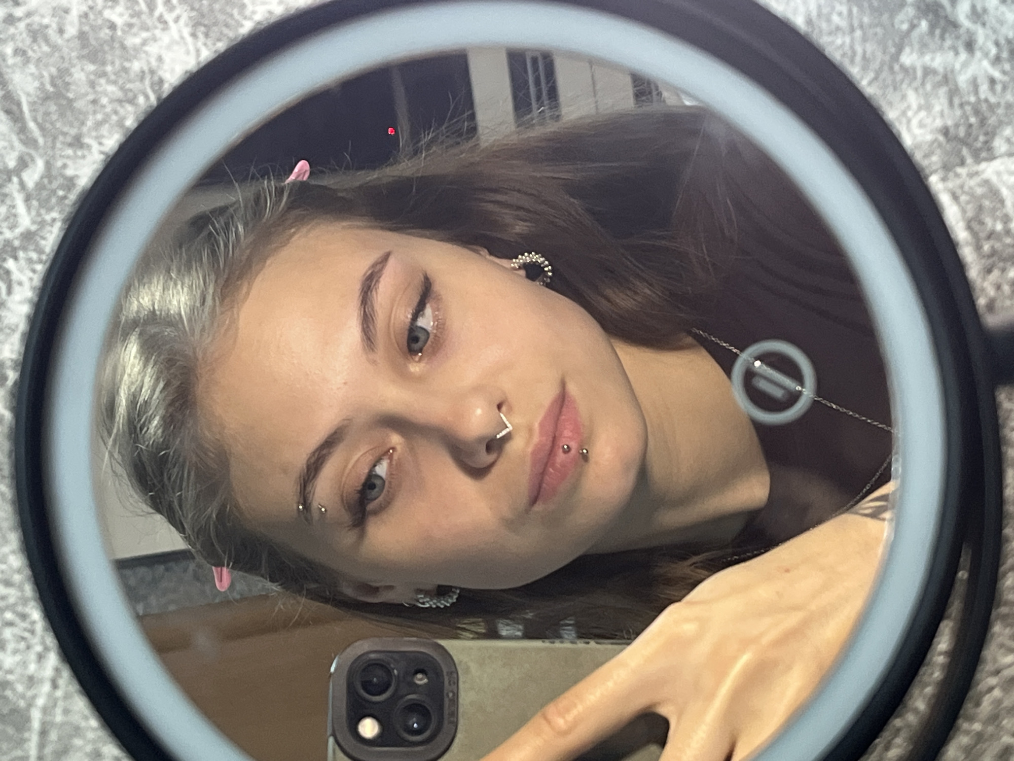 A young woman is taking a selfie in a round mirror. The mirror is illuminated by a ring light. The woman has light skin, light brown hair, and blue eyes. She has multiple facial piercings, including an eyebrow piercing, a nose piercing, and lip piercings. She is wearing earrings and a necklace. The phone she is using has multiple cameras. The location in the city of Фоки, Russia cannot be determined from the image.