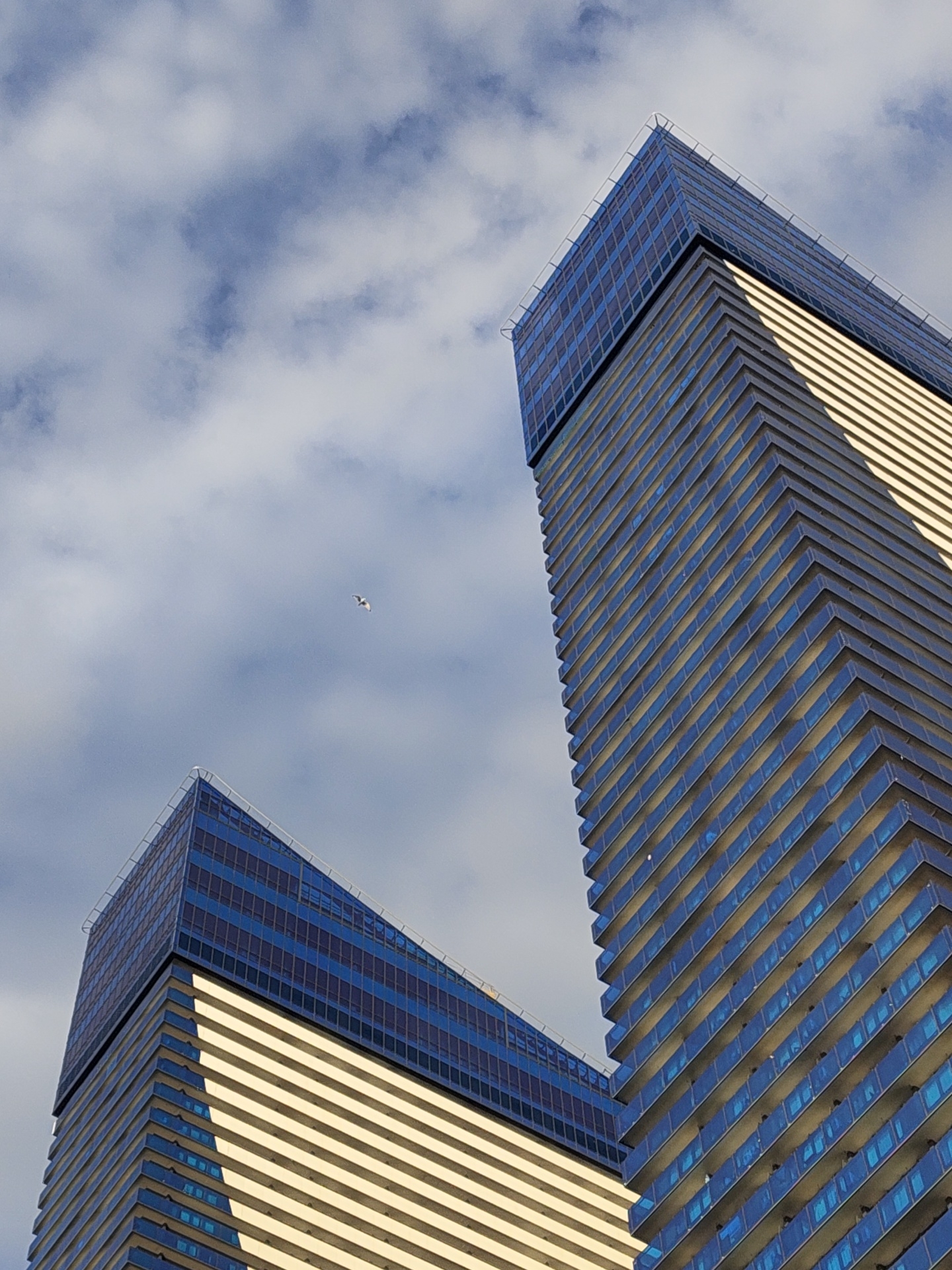 Two modern skyscrapers with angled, multi-faceted facades of glass and light-colored horizontal bands rise towards a cloudy sky. The taller skyscraper on the right is closer to the viewer and its facade shows diagonal lines created by the building's structure. The skyscraper on the left is further away and appears to be slightly shorter. A small, white bird is visible flying in the sky between the two buildings. The image is a low-angle shot, emphasizing the height and architectural design of the buildings, and the clear blue sky with scattered clouds. The city of Batumi, Georgia, is known for its contemporary architecture, particularly its seaside skyscrapers, which often feature innovative designs and reflective glass facades like those depicted.