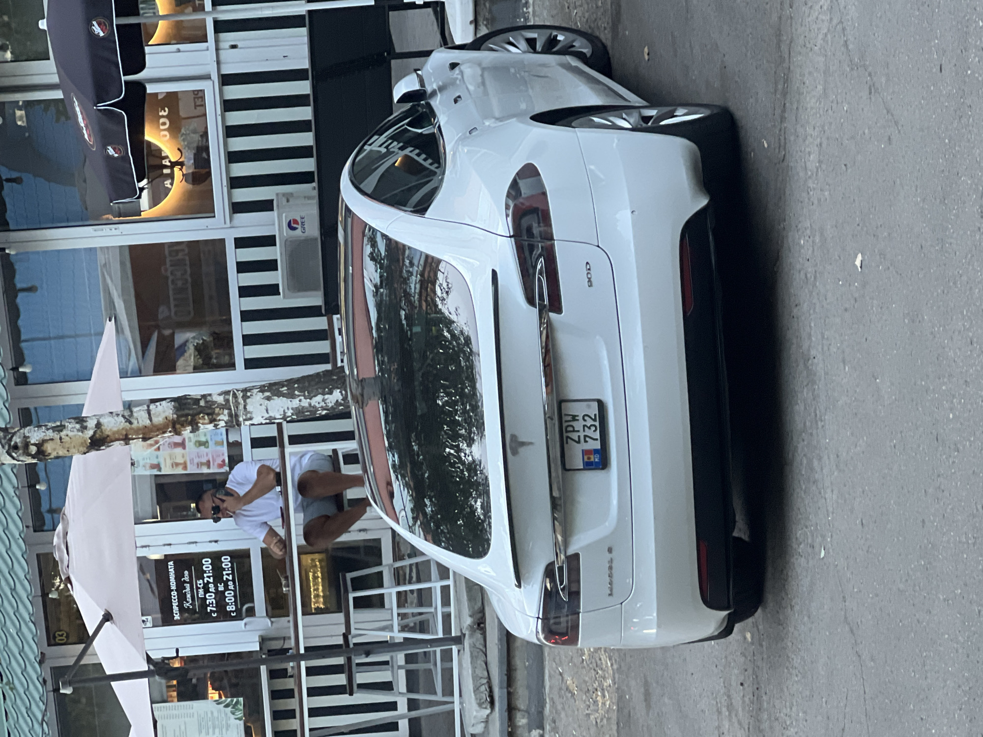 A white Tesla Model S is parked on an asphalt surface in front of a building with black and white striped siding. The car's rear is visible, showing a license plate with the characters "ZPW 732" and "MD" indicating Moldova. To the left of the car, a man in a white t-shirt and gray shorts is seated at a table outside a cafe, smoking and talking on a mobile phone. He is positioned under a large, pale pink umbrella, with a tree trunk to his right. Behind the man, a sign in Russian lists cafe operating hours and offerings. The building has large windows reflecting the surroundings, and an air conditioning unit is mounted on the exterior wall. The scene appears to be in the city of Bender, Moldova.