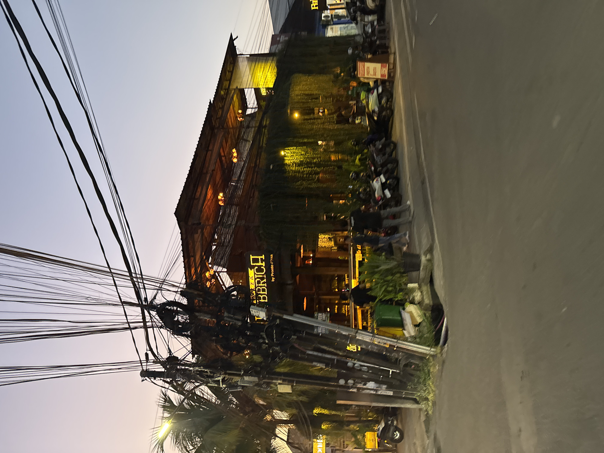 In North Kuta, Indonesia, at dusk, a two-story building with an open-air upper level is illuminated. The building is covered in hanging greenery on its facade. A prominent sign reads "UBBRICH by Pronto Pizza." A dense network of electrical wires hangs from poles in the foreground, obscuring parts of the building. Several motorbikes are parked in front of the establishment. The street in front of the building is paved, and a sidewalk is visible. People are seen standing and walking near the entrance. The sky is a gradient of pale orange and blue, indicating the end of the day.