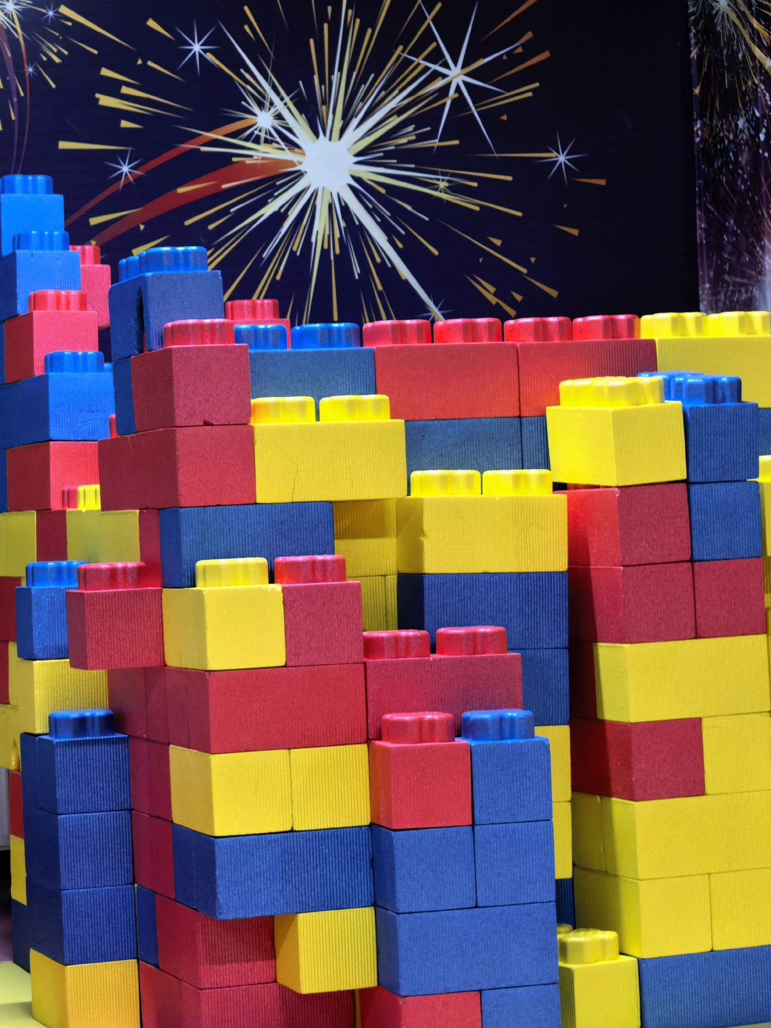 A close-up shows a tall, complex structure made of large, colorful building blocks, predominantly red, yellow, and blue. The blocks are arranged in a terraced or step-like formation. In the background, there is a backdrop featuring a dark blue or purple color scheme with what appears to be a stylized representation of fireworks or starbursts. The building block structure is on a surface that appears to be a yellow or light-colored table. There is no sign of human activity in the photograph. Without other information, no information exists to identify the location in Sterlitamak, Russia.