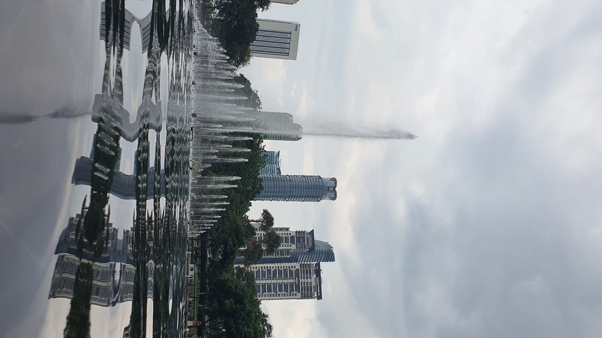 The image captures a wide shot of the Kuala Lumpur City Centre Park with its expansive water features. In the foreground, a large, dark pool of water reflects the surrounding cityscape and the elaborate fountain display. The fountain itself is a prominent feature, with numerous jets of water arcing upwards, creating a dynamic visual. In the background, a line of modern skyscrapers rises against a cloudy sky, indicative of a bustling urban environment. One building, marked with the PNB logo, stands out with its distinct blue and white facade. Lush green trees are interspersed between the buildings and along the park's edge, providing a natural contrast to the man-made structures. A few people are visible in the background, suggesting the park is a public space enjoyed by visitors. The overall impression is one of a well-maintained urban park offering a serene escape within the heart of Kuala Lumpur.