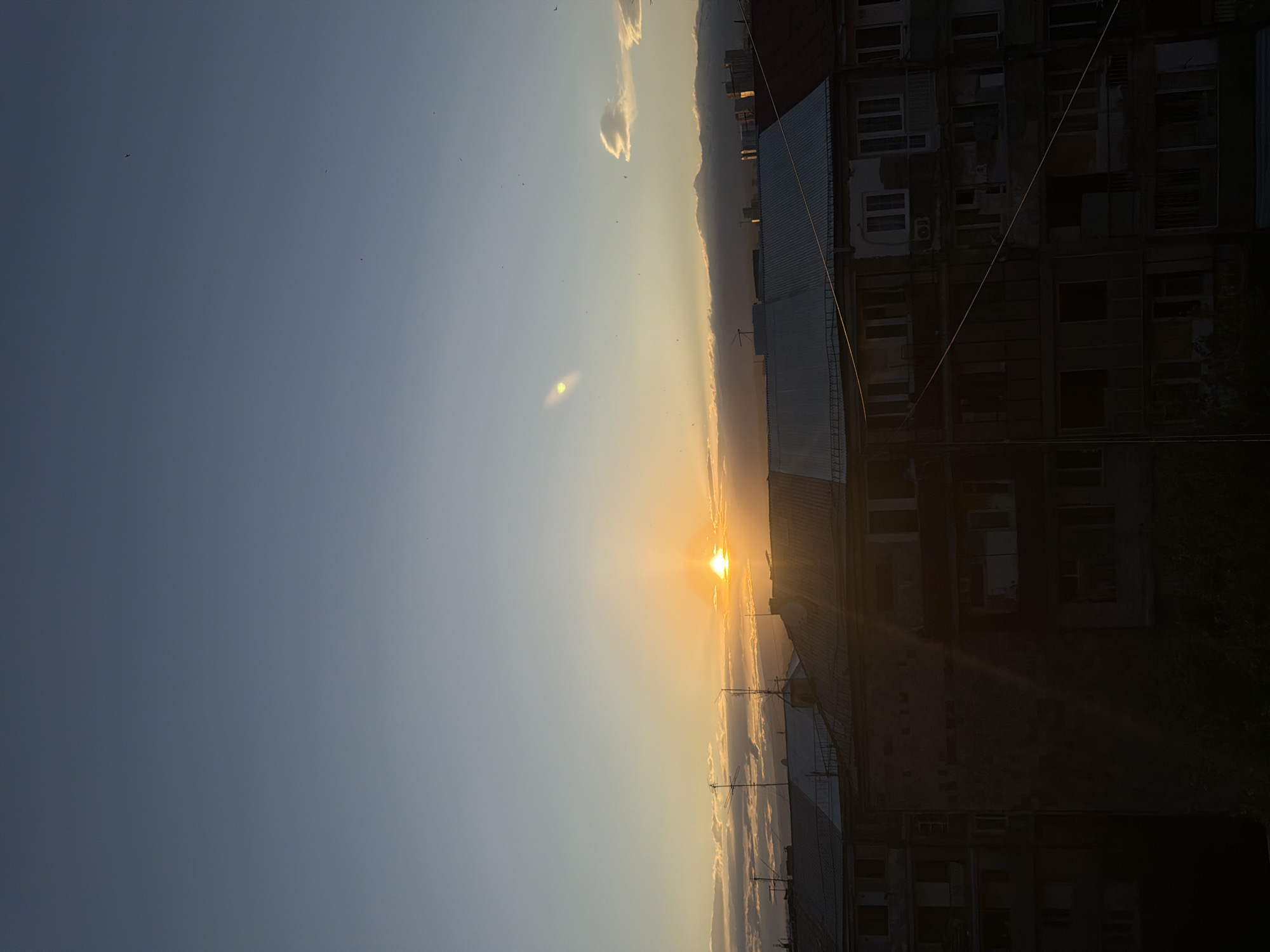 The image depicts a sunset in Yerevan, Armenia. The sun is setting behind mountains, with a city skyline visible in the distance. In the foreground, there are buildings, including an apartment complex. The sky is a gradient of blue and orange, with a few small clouds. There are a few birds flying in the sky.