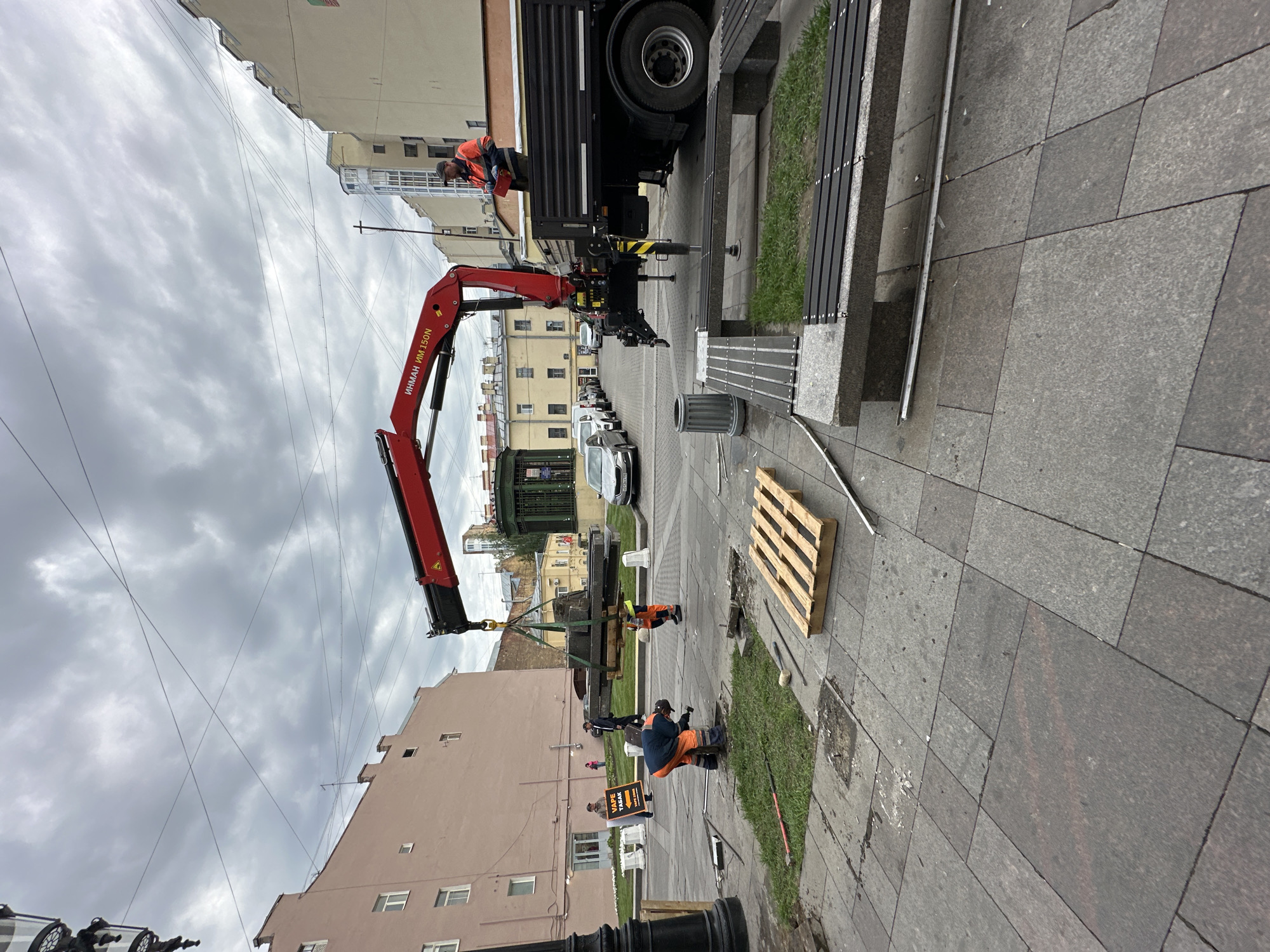 The image captures a streetscape in Saint Petersburg, Russia, featuring a crane truck and workers engaged in street maintenance or construction activities. A red "ИНМАН ИМ 150N" crane is centrally positioned, extending its arm to lift a section of the pavement, likely part of a landscaping or repair project. A pallet rests on the sidewalk near a gap where paving stones appear to have been removed.
Two workers in orange vests are in view; one is operating near the lifted section while another is seated, seemingly engaged in preliminary work or supervision. A person holds a sign reading "VAPE TABAK"
The backdrop includes typical urban elements such as buildings of varied heights and architectural styles, a row of parked cars along the street, and overhead power lines. The sky is overcast, contributing to a muted color palette. A bench is placed on the sidewalk.