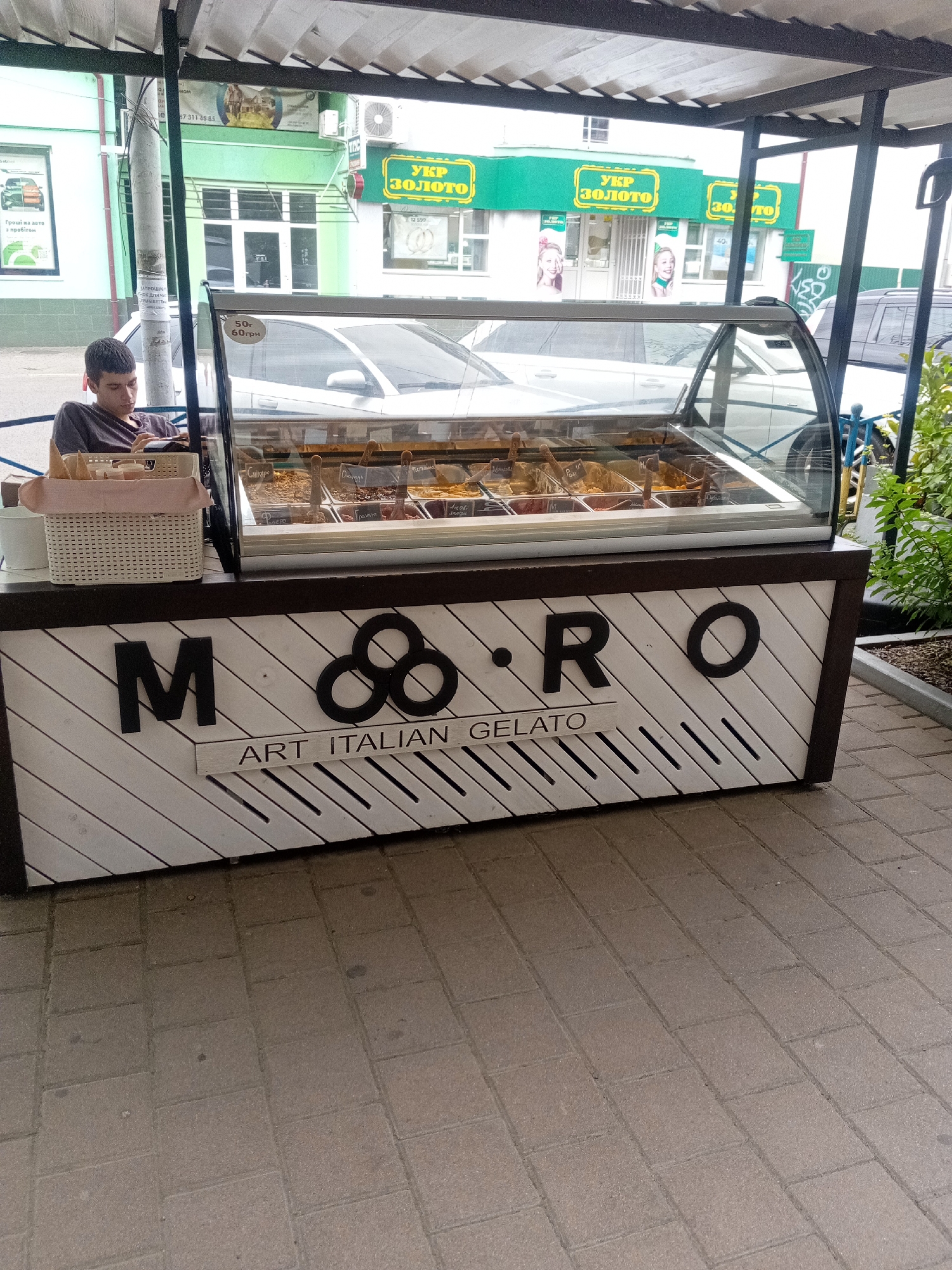 A young man sits at a gelato stand in Tiachiv, Ukraine, engrossed in a book. The stand, featuring a glass display case filled with various gelato flavors, is decorated with the words "M & O ART ITALIAN GELATO" in bold black lettering. Behind the stand, a row of buildings line the street, including a store with a prominent green sign reading "УКР ЗОЛОТО" (UKR GOLD). The pavement in front of the stand is made of rectangular pavers. The overall scene suggests a typical day at a local business.