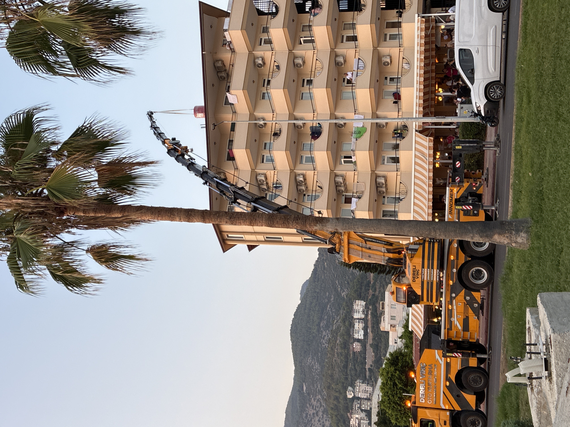 In Alanya, Turkey, a large yellow crane is positioned on a street, its boom extended towards a multi-story apartment building. The building, painted a light peach color, has numerous balconies with metal railings and is adorned with many air conditioning units and laundry hanging out to dry. A red cylindrical object is suspended from the crane's hook, nearing the rooftop. Palm trees are visible in the foreground and framing the scene. In the background, rolling green hills dotted with white buildings rise under a pale sky. A white van is parked on the street in front of the apartment building, and a few people are visible on the balconies of the building and at outdoor tables in front of a ground-floor establishment. The overall scene suggests ongoing construction or a delivery operation in a coastal city.