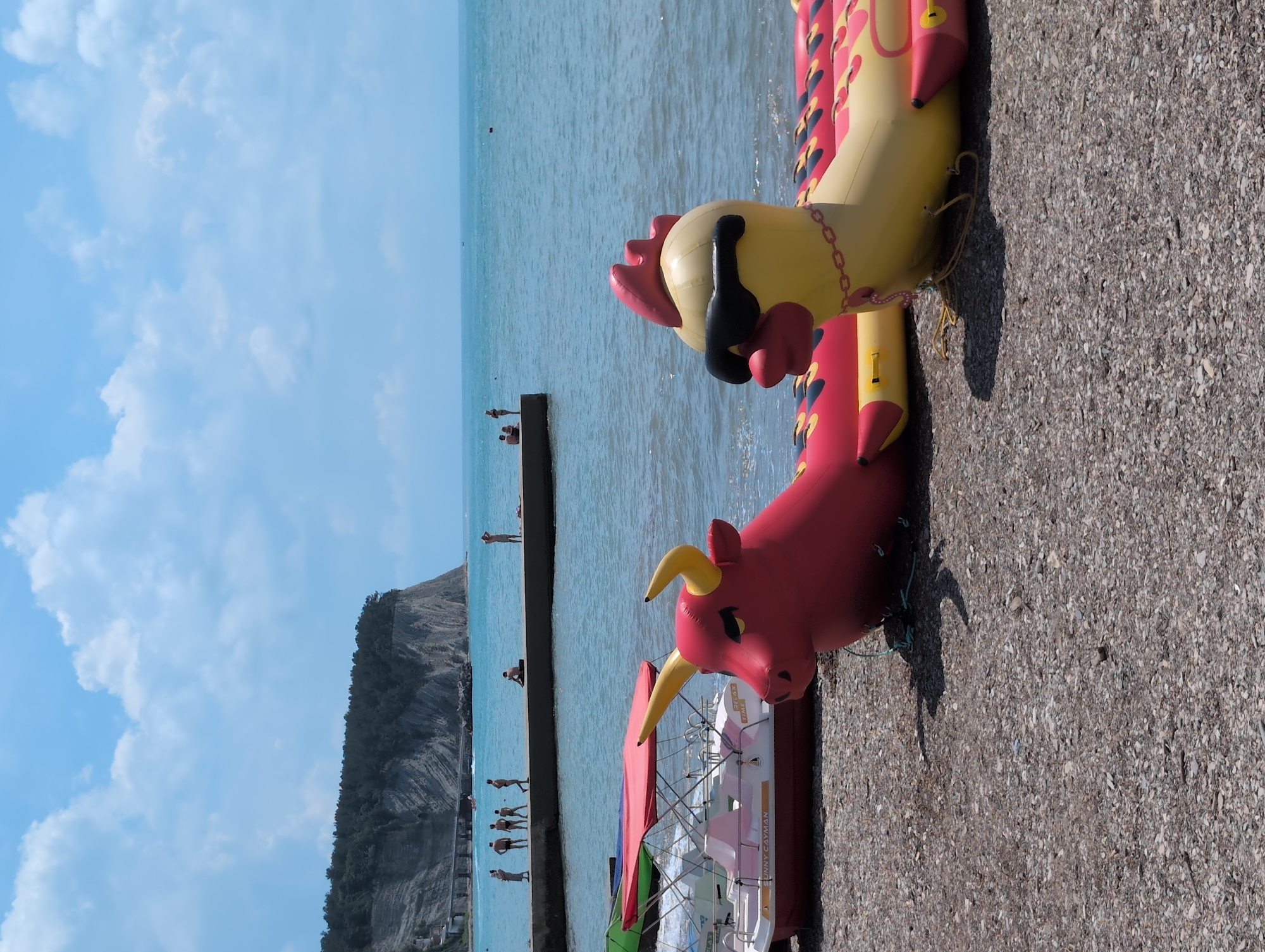 On a pebble beach in Divnomorskoye, Russia, two inflatable water toys, a red bull and a yellow chicken wearing sunglasses, lie in the foreground. To the left, a white boat with a red canopy is docked. In the background, people walk along a concrete pier that extends into the turquoise sea. A large, tree-covered cliff rises on the left side of the scene, and a clear blue sky with scattered white clouds stretches above.
