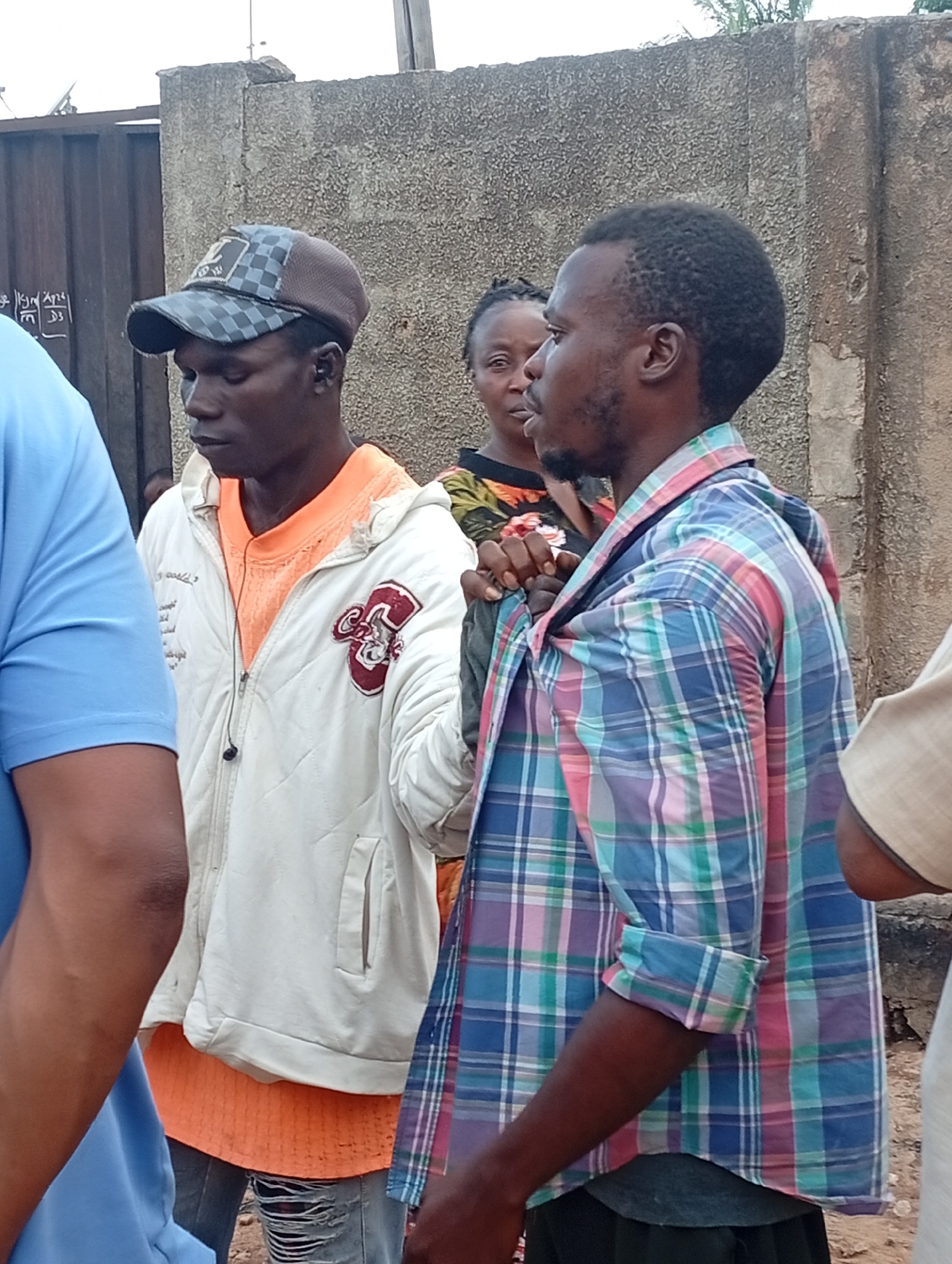 In Sabon Tasha, Nigeria, a group of people are standing outdoors. A man is wearing a plaid shirt. Another man wears a white jacket over an orange shirt, with ripped jeans and a baseball cap. A woman stands in the background. The background shows a concrete wall and a metal gate.