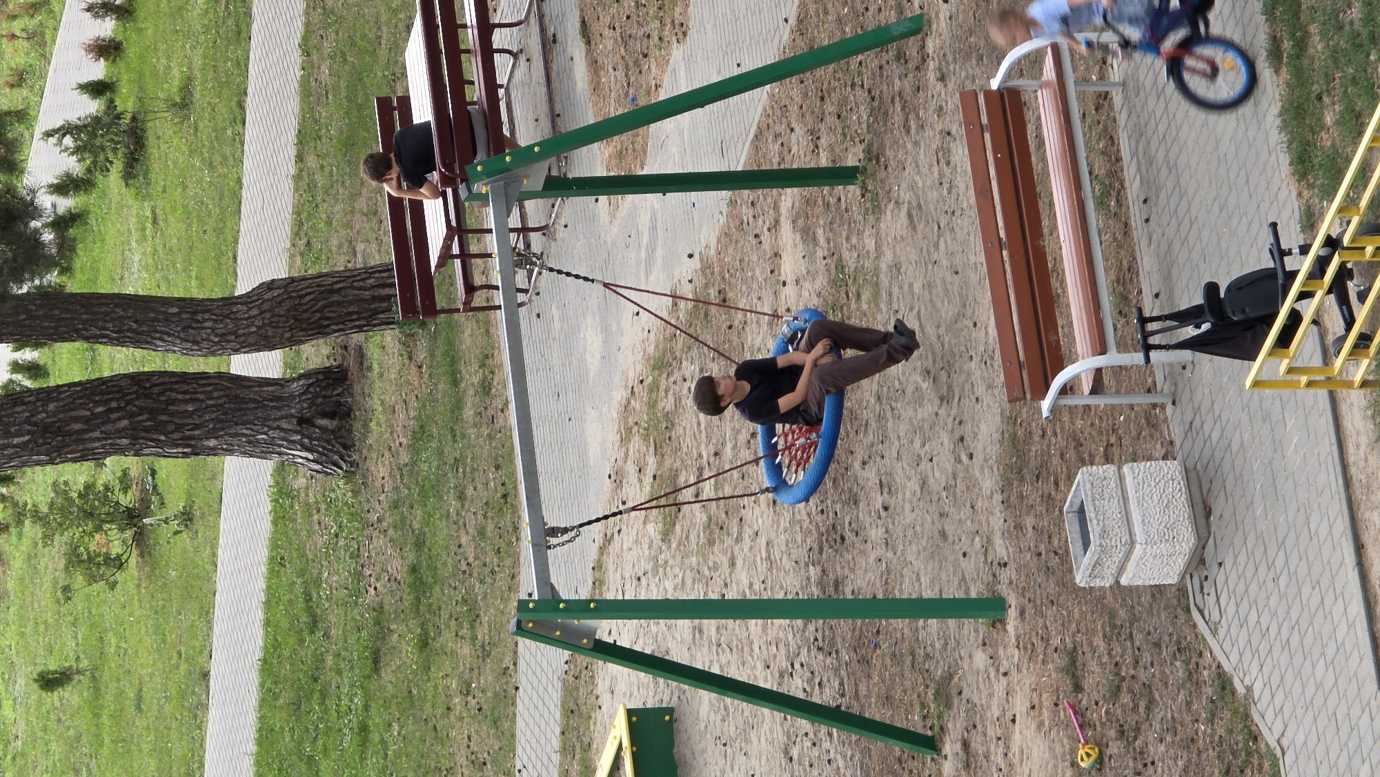 A high-angle, medium shot captures a playground scene with a focus on a young boy on a swing. The boy, wearing a dark t-shirt and brown pants, is seated on a round, blue net swing suspended from a metal frame with green support poles. He looks to his right, his gaze directed slightly downwards. The swing set is situated on a sandy surface, with scattered pinecones and dried grass. In the background, two large pine trees stand prominently. To the left of the swing, a portion of a yellow and green sandbox structure is visible. Further back, a concrete path winds through a grassy area with some small trees and shrubs.
To the right of the swing, a wooden park bench with a metal frame is positioned on a paved area. Behind this bench, another, longer wooden bench with a similar frame is occupied by a young person in a black t-shirt, facing away from the camera and resting their chin on their hand. This bench overlooks a paved area with a row of small trees.
In the foreground, to the right of the frame, a child on a bicycle with a blue rim on the front wheel is in motion, partially blurred by speed. In front of the longer bench, a concrete planter with a textured surface sits on the paving stones. A yellow plastic toy, resembling a steering wheel, is on the ground to the left of the planter.
The image depicts a public outdoor space, likely a park or playground, characterized by natural elements like trees and grass, as well as recreational equipment and urban infrastructure such as benches and paved pathways. The scene suggests a casual, everyday setting.