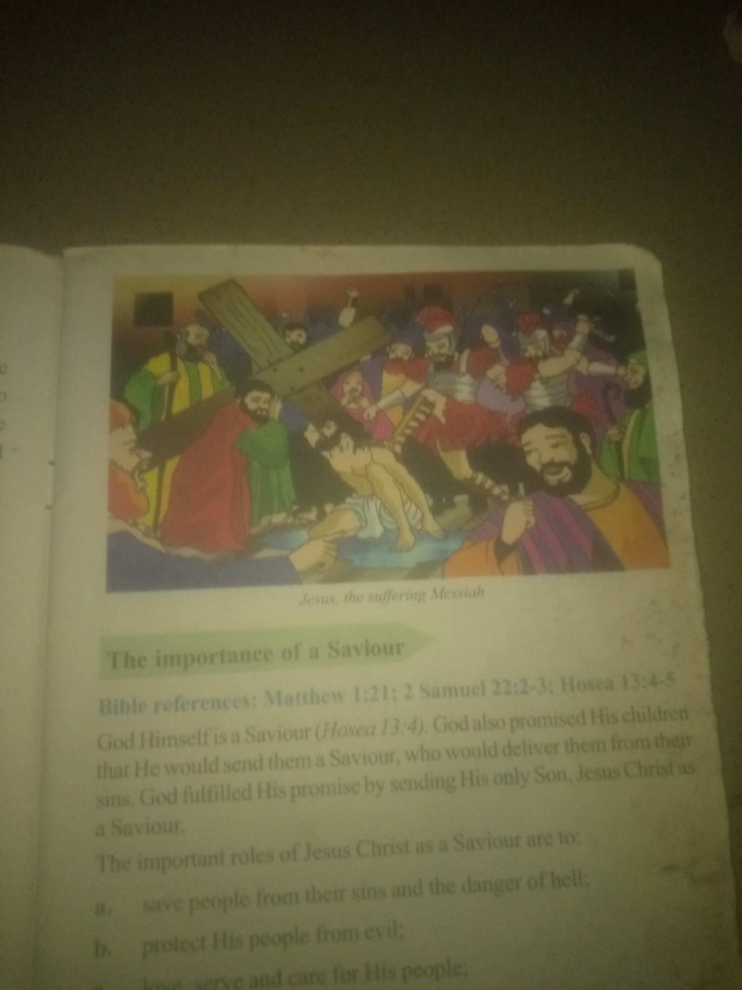 The image is a photograph of an open book. The book displays an illustration depicting Jesus carrying the cross, surrounded by a crowd of people. Below the illustration, there is text that reads "Jesus, the suffering Messiah." Further down, a green banner contains the title "The importance of a Saviour." Beneath this title, several lines of text discuss Bible references and the roles of Jesus Christ as a Savior. The book appears to be open on a surface in a dimly lit environment, possibly a room. There is no visible indication of a specific city or location within Nigeria.