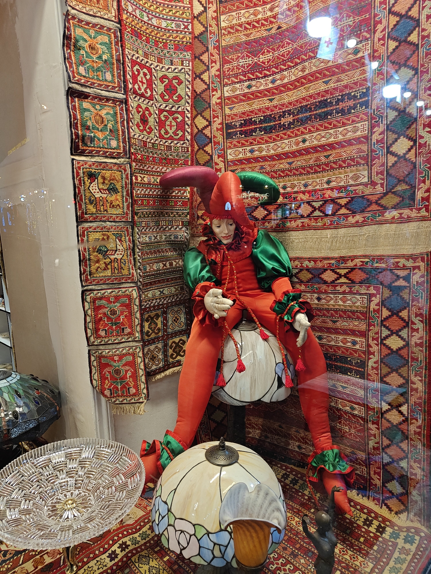 This vertical image depicts an ornate display, likely within a shop window or an indoor retail setting. The central subject is a whimsical jester or harlequin doll, seated and dressed in a vibrant red and emerald green costume. The doll features a pointed jester's cap, ruffled collar and cuffs, and holds red tasseled cords. Its face is pale with delicate, doll-like features.
The background is dominated by a large, intricately patterned rug or tapestry, rich with geometric designs and stripes in shades of red, gold, black, green, and blue. To the left, a vertical series of smaller, square textile pieces, possibly small tapestries or pillow covers, hang against a white wall. These pieces feature distinct patterns, including stylized eagles or birds and roosters, in similar vibrant color palettes of green, red, and yellow.
In the foreground, several decorative items are showcased. A large, clear, cut-glass bowl with an intricate woven or starburst pattern rests on a stand to the left. Below the jester, a prominent Tiffany-style lamp with a stained-glass shade displays floral or abstract motifs in blues, greens, and creams. A smaller, metallic lamp element is visible nearby. Towards the bottom right, a dark statuette of a figure with raised arms is positioned next to the Tiffany lamp.
The entire scene is observed through glass, as indicated by numerous bright, circular reflections of lights scattered across the surface, particularly prominent on the large rug. These reflections suggest the display is either in a well-lit interior or viewed from outside at night. The overall impression is one of rich craftsmanship and decorative artistry, consistent with traditional wares found in places like Tbilisi, Georgia. Faint, illegible text appears in the reflections on the far left.