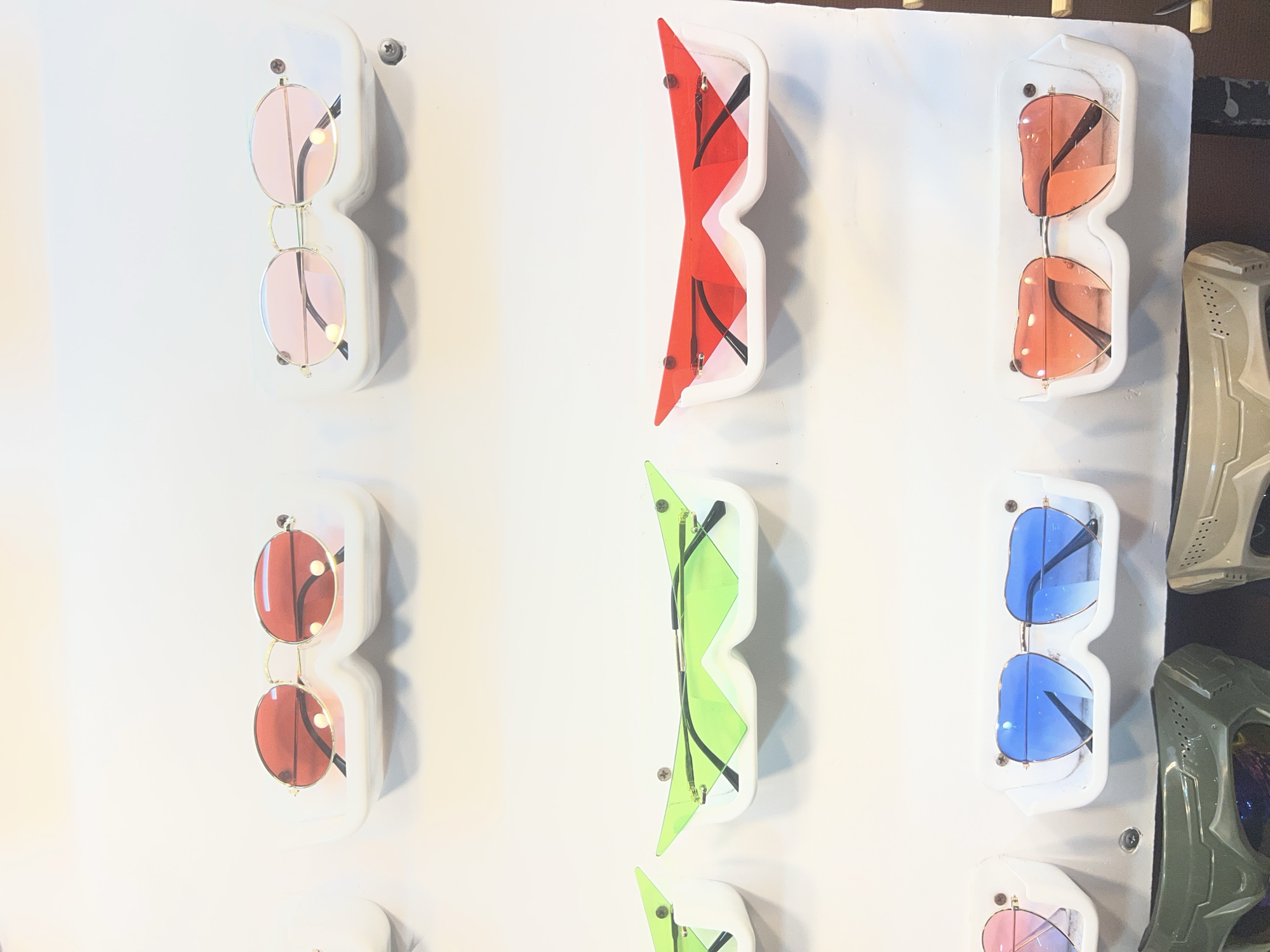 The image shows a display of various sunglasses on a white background. The sunglasses are arranged in rows and are held in individual white holders. In the top row, there are two pairs of round sunglasses, one with red lenses and the other with pink lenses. The middle row features two pairs of uniquely shaped sunglasses, one with bright green lenses and the other with red lenses, both with sharp, angular designs. The bottom row displays two pairs of heart-shaped sunglasses, one with blue lenses and the other with orange-pink gradient lenses. Below the sunglasses display, parts of what appear to be protective masks or gear are visible, one in olive green and another in a light tan color. The overall environment appears to be a retail setting, possibly a shop or market stall, in ตำบลเกาะพะงัน, Thailand. There are no people visible in the image.