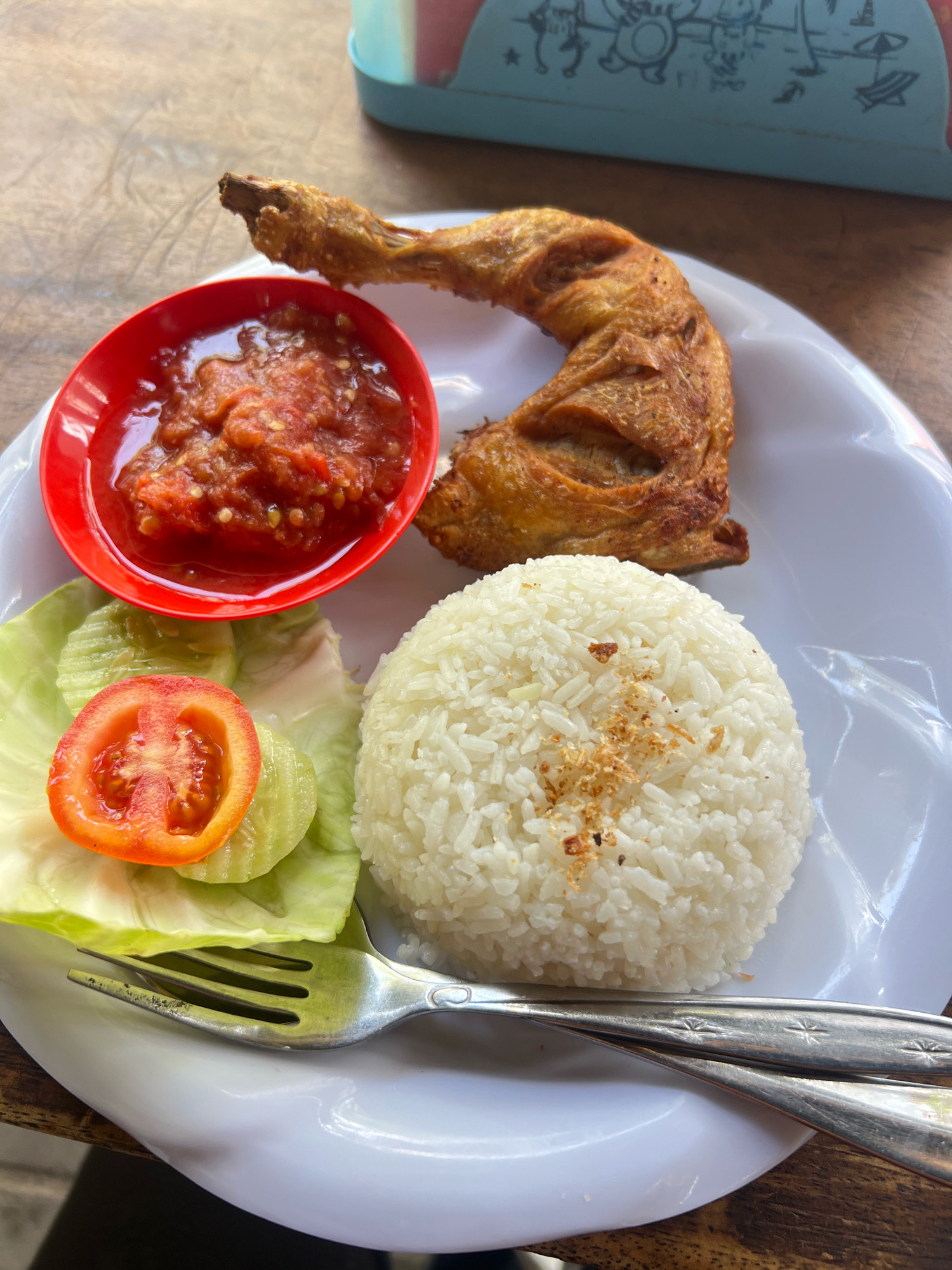 The image shows a plate of food, likely from a restaurant in Kuta, Indonesia. The plate contains a piece of fried chicken leg, a mound of white rice, a small red bowl of chili sauce, and a side of fresh vegetables including lettuce, sliced cucumbers, and a slice of tomato. A fork is placed on the plate. The food is arranged on a white plate, which sits on a wooden table. In the background, a colorful plastic container with cartoon drawings is visible. The lighting suggests an outdoor or well-lit indoor setting.