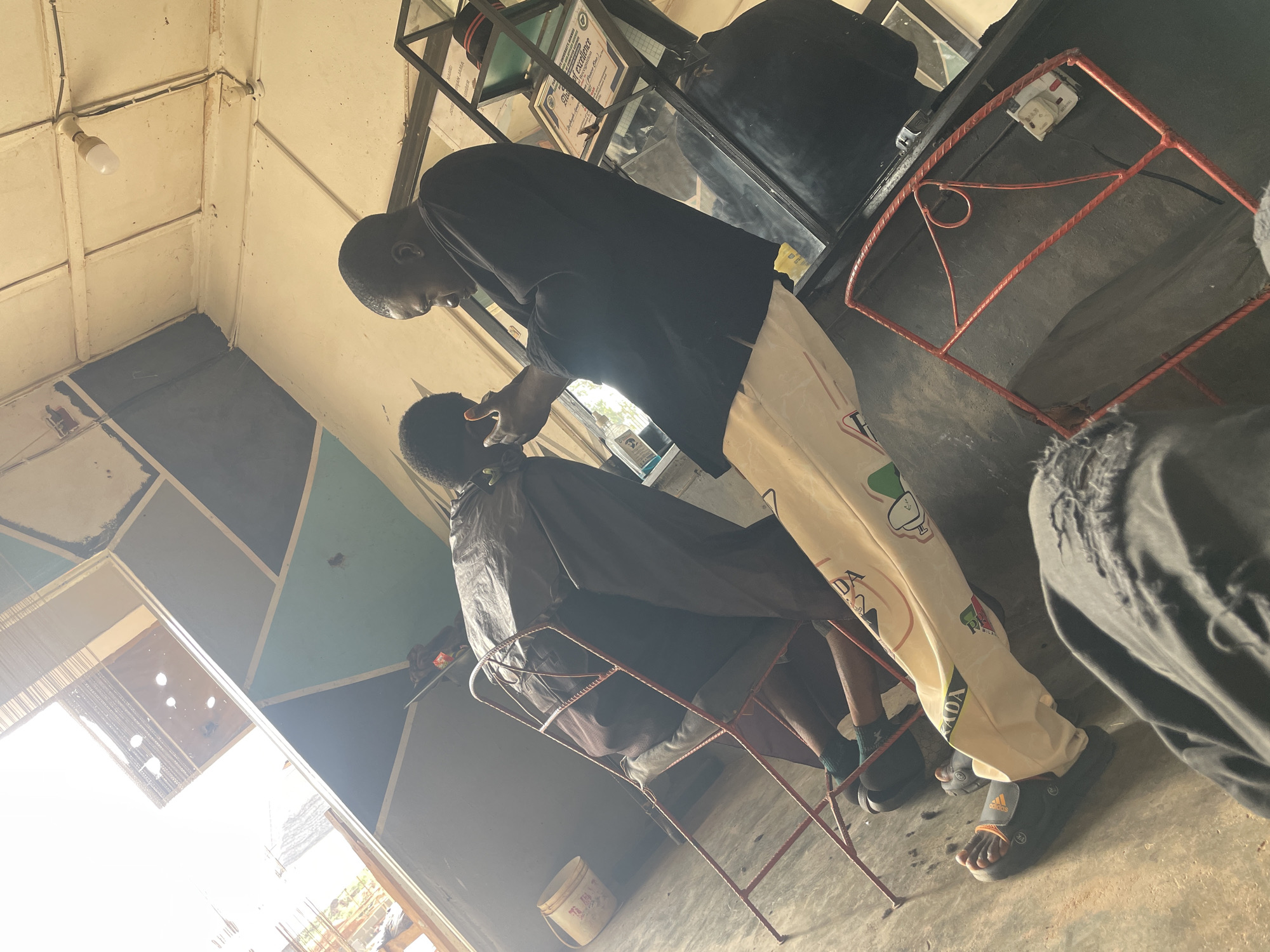 A barber in Durumi, Nigeria, is giving a haircut to a customer. The barber is wearing a black t-shirt and light colored patterned pants. The customer is covered with a black cape. They are in a simple barbershop.