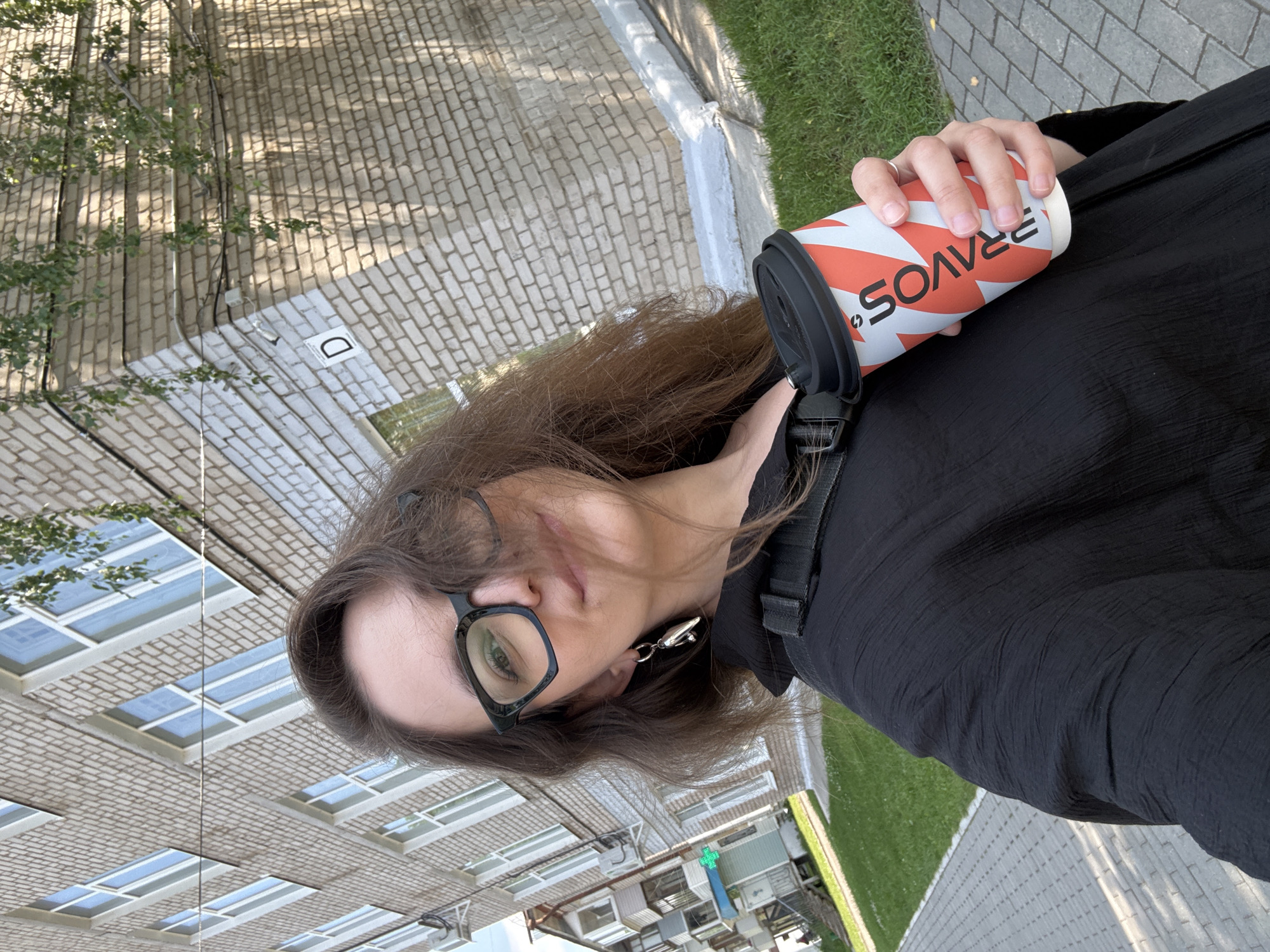 A selfie is taken by a young Caucasian woman with long, dark brown hair and black-rimmed glasses. She is wearing a black, textured top. In her right hand, she holds a disposable coffee cup with an orange and white striped pattern and the word "PRAVOS" visible. She is standing outdoors on a paved walkway next to a green lawn. Behind her is a light-colored brick building with multiple windows and some overhanging tree branches. The setting appears to be in the city of Chaikovsky, Russia, suggested by the architectural style of the building and the visible signage on the building, which is typical of Russian residential buildings. The lighting indicates it is daytime, with sunlight casting shadows on the building.